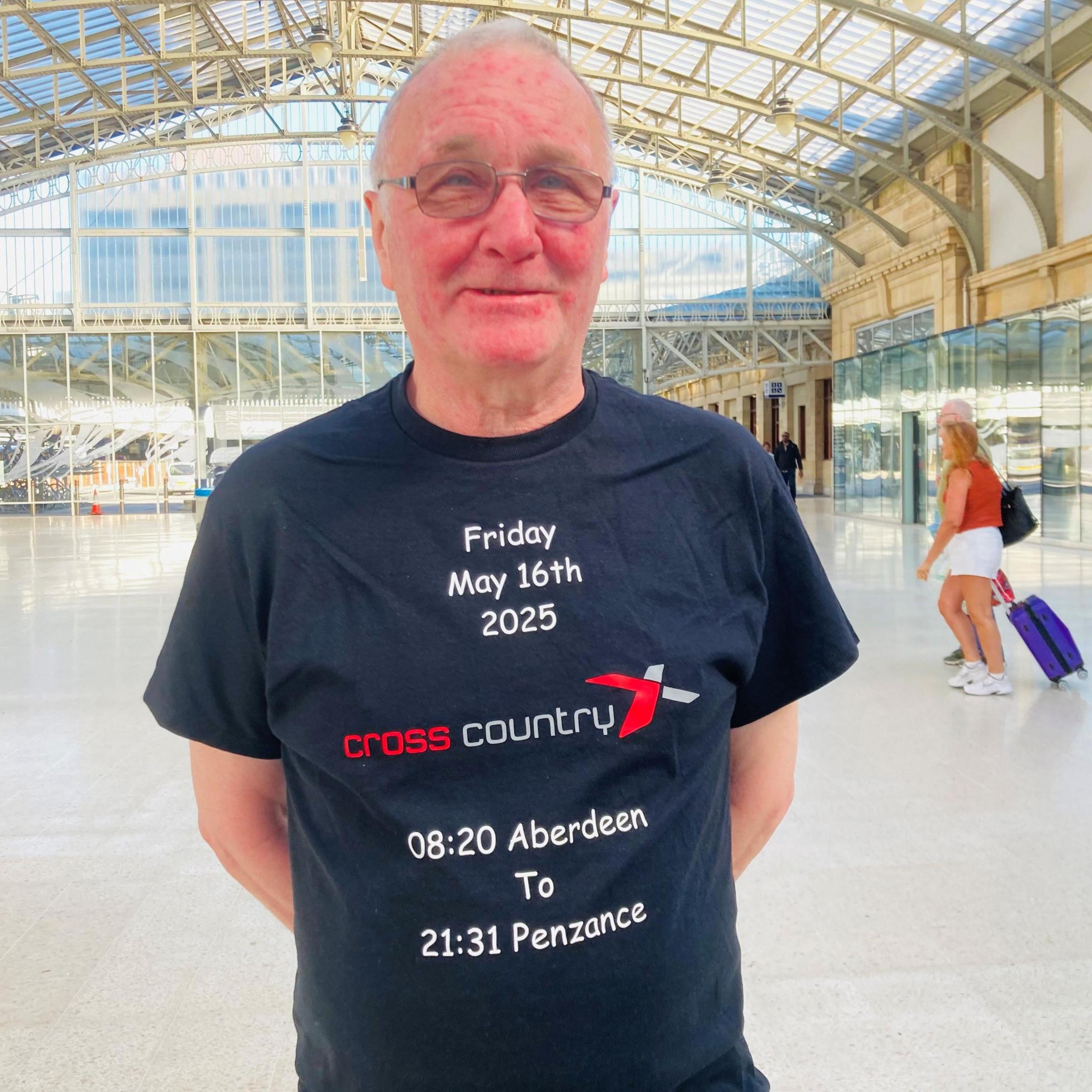 Man - John Labrow - smiling at camera in station, wearing a t-shirt which says 'Friday May 16th 2025 / CrossCountry / 08:20 Aberdeen to 21:31 Penzance'.