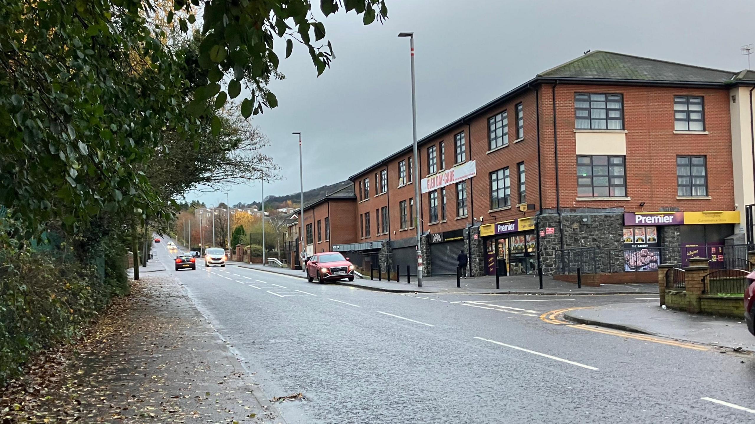 A street with a modern red brick building and shopfronts at the bottom. Ove red car is parked on the pavement and a couple of cars are driving down the street. 