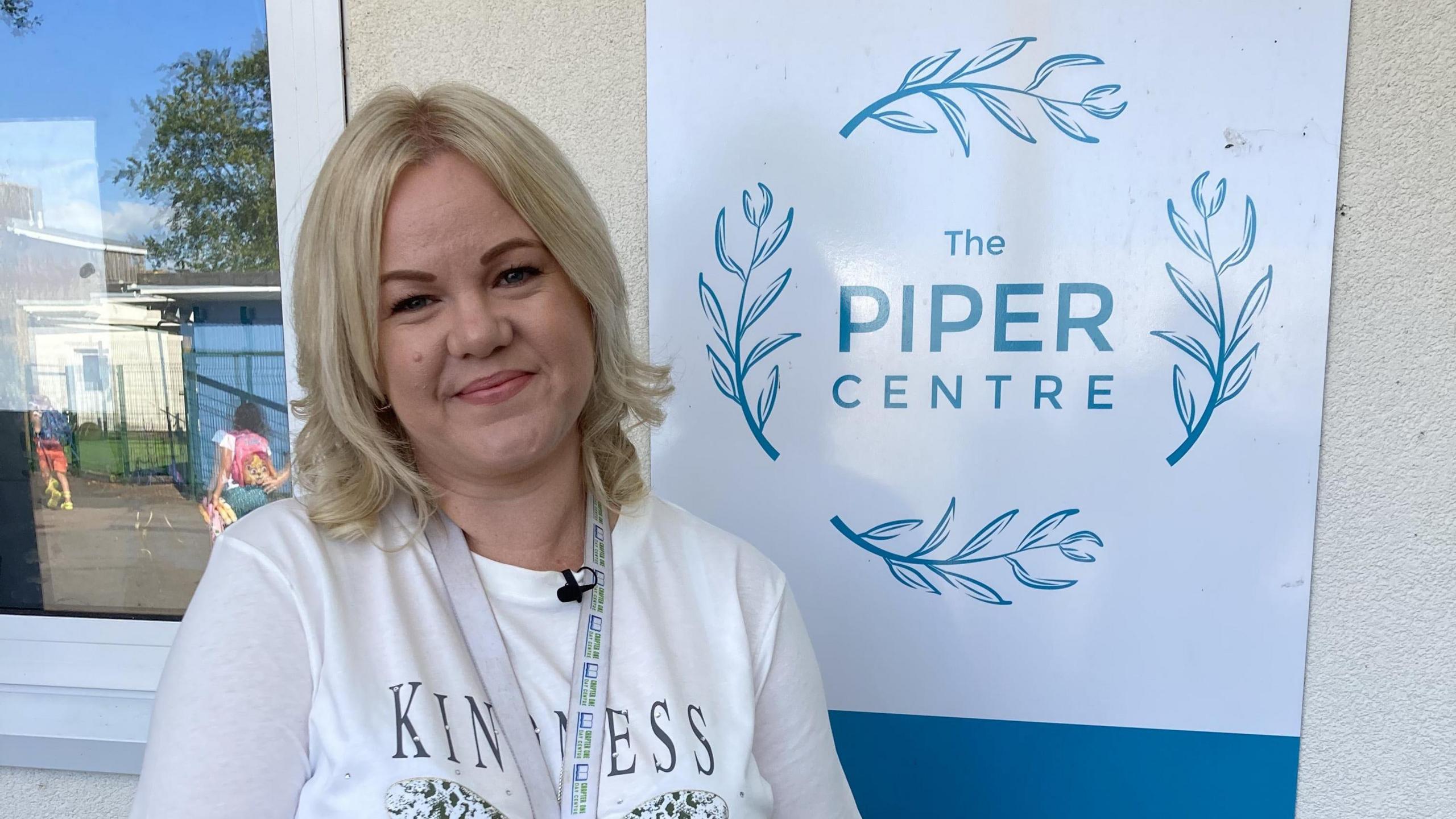 A woman called Shell Potter stood in front of a sign which reads, 'The Piper Centre'. She is smiling and is wearing a white long-sleeved top with the word "kindness" on it