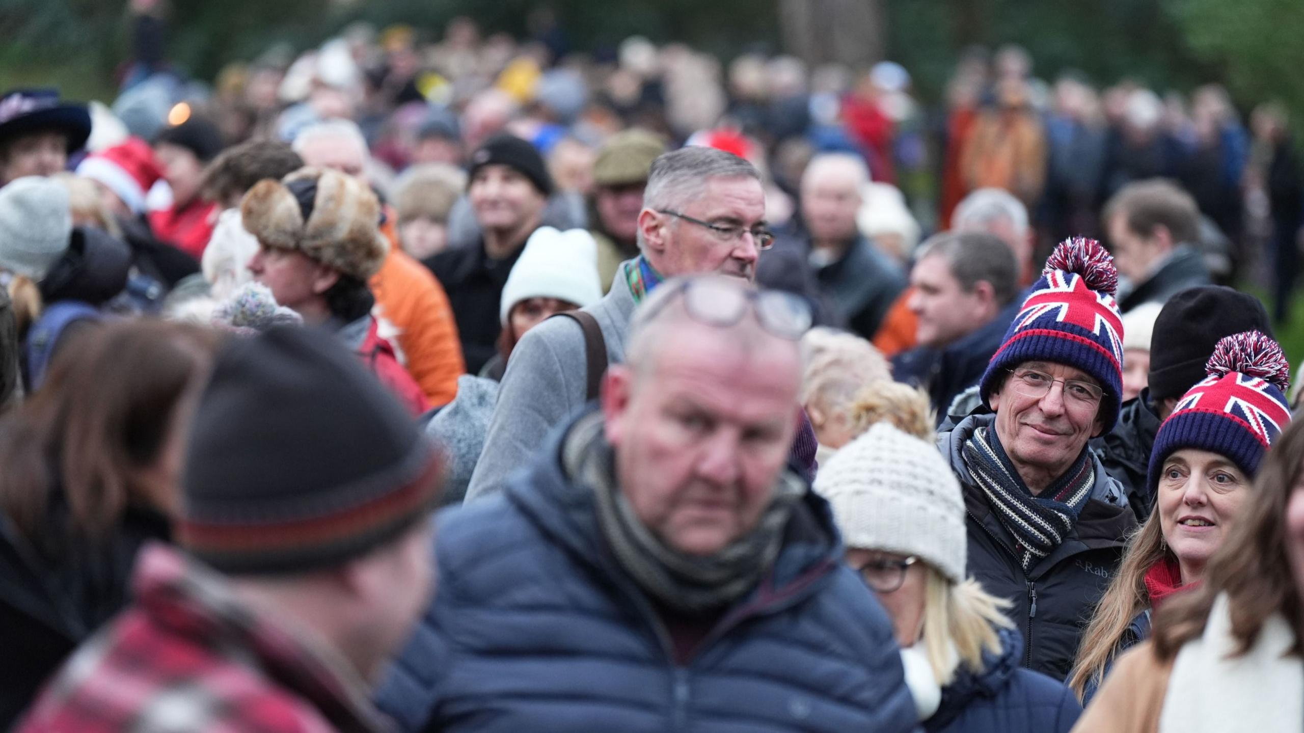 Members of the public gather ahead of the Christmas Day morning church service attended by the royal family at St Mary Magdalene Church in Sandringham, Norfolk. People are wearing puffer jackets, beanies and chat among themselves. 