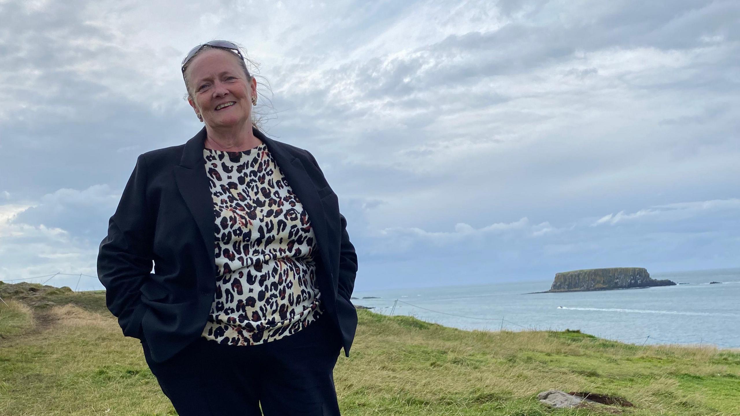 Suzanne Buckner stands on a grassy clifftop with the sea and a small island in the background. She wears a black-and-white blouse, dark trousers and a dark jacket, with sunglasses pushed back over her brown hair.
