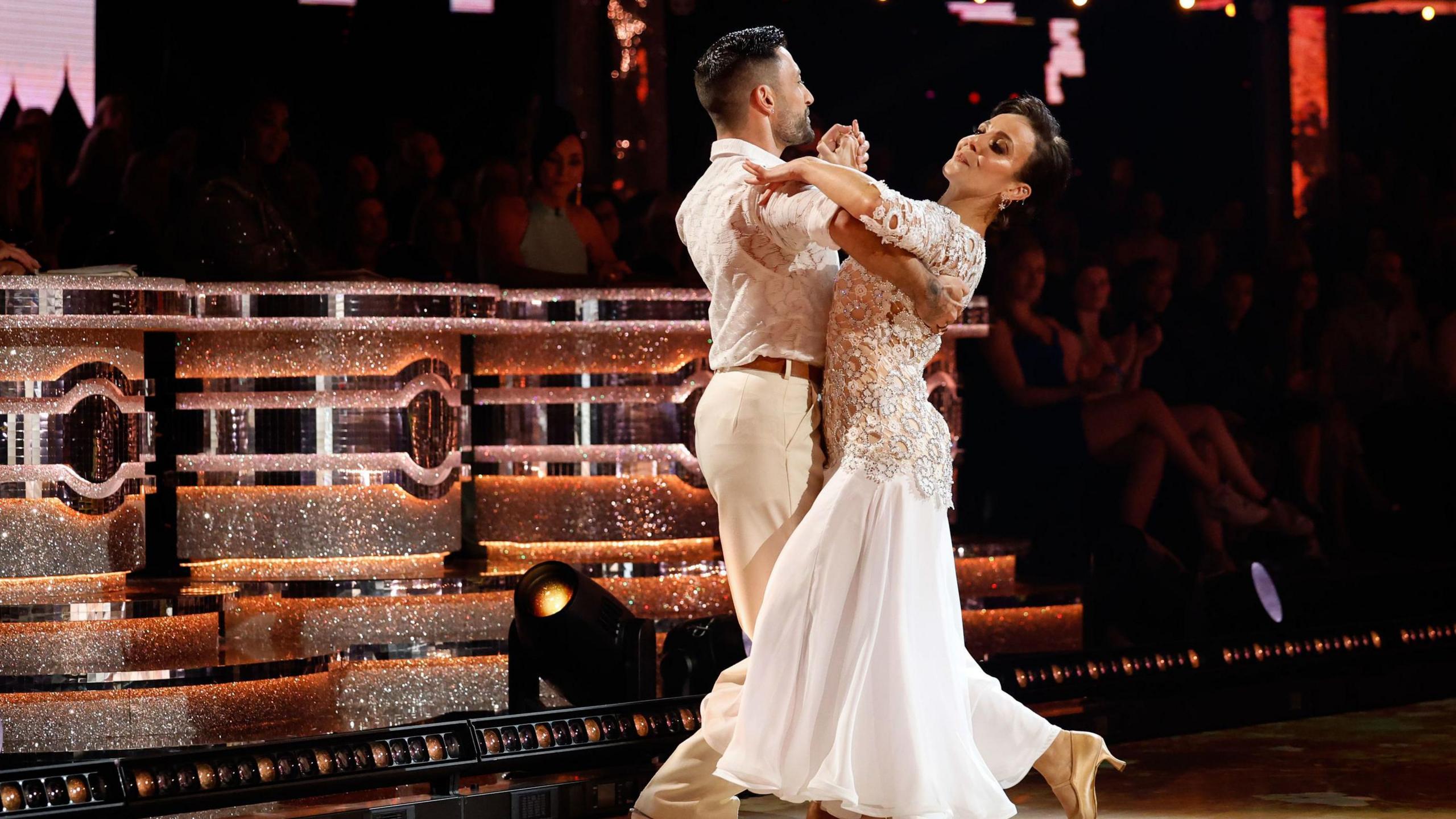 Both dressed in white and cream colours, Giovanni Pernice and Amanda Abbington dance on the Strictly stage. The judges' panel can be seen behind them as well as some audience members although the light is focussed on the couple. 