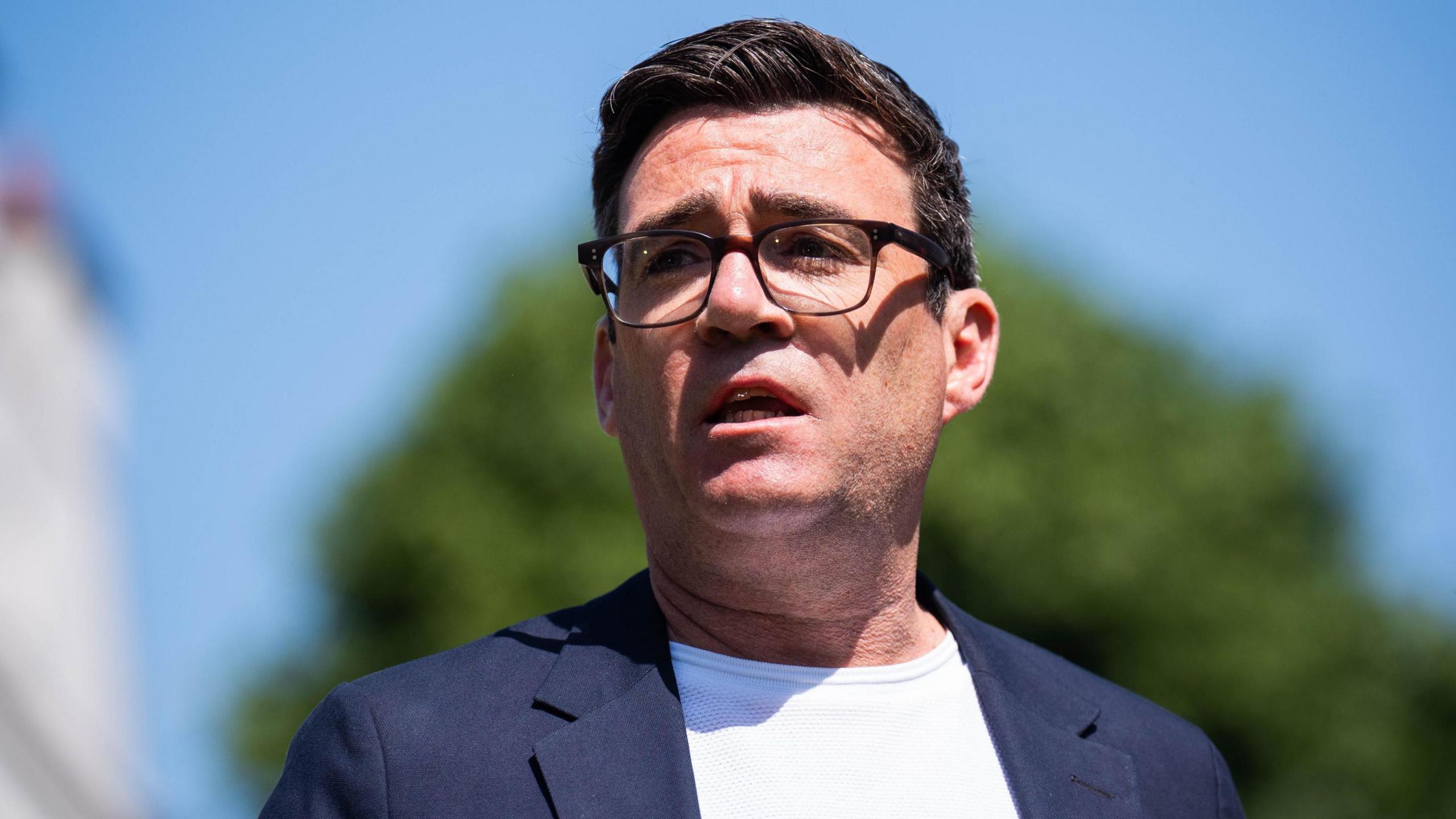 Image shows Andy Burnham, the mayor of Greater Manchester, wearing glasses, a while t-shirt and navy blue suit jacket. 