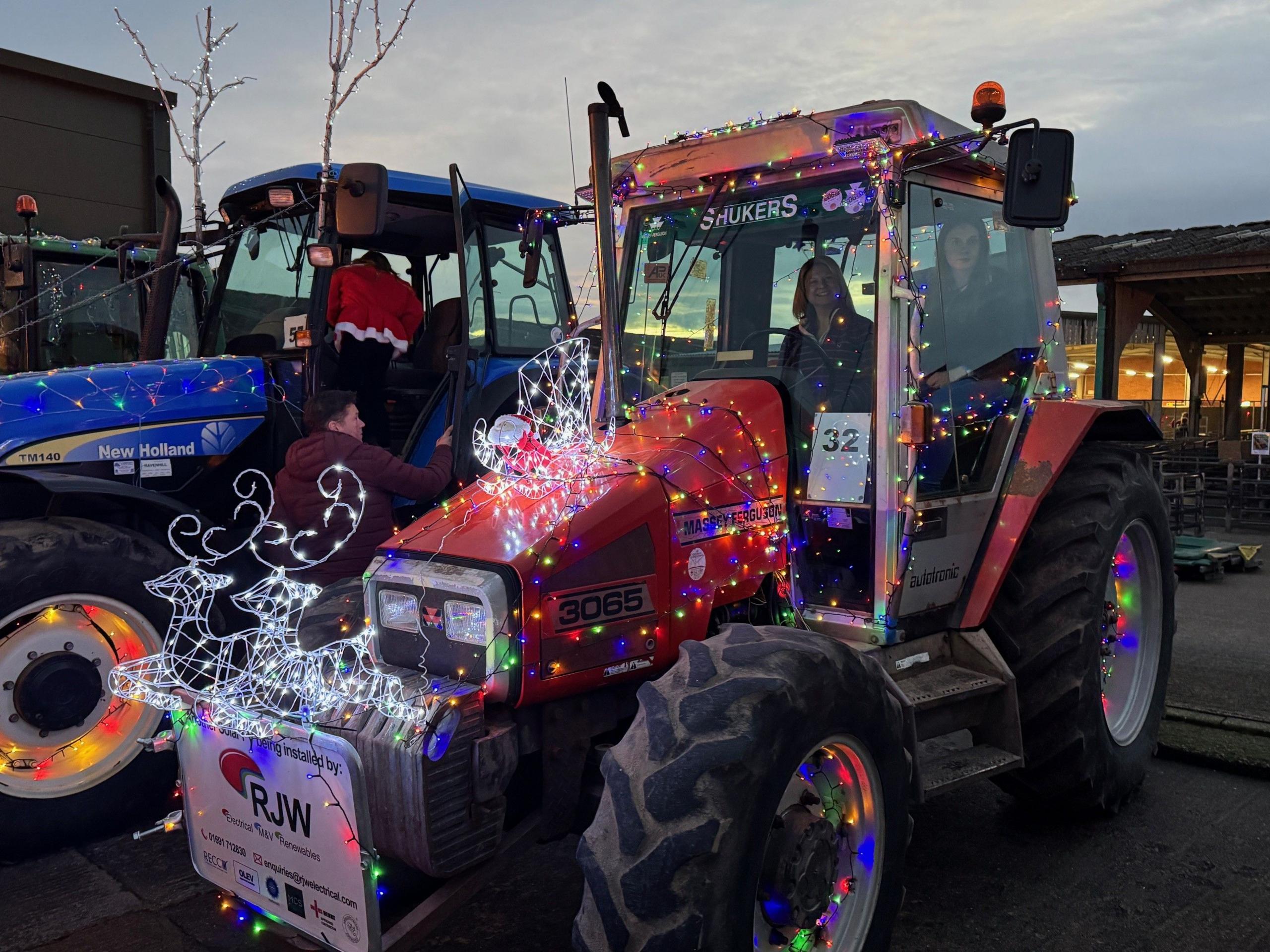 A red tractor and a blue tractor parked next to each other, covered in multi-coloured Christmas lights. The red tractor is nearest the camera and has two white reindeer sculptures made of Christmas lights. Two women are in the tractor on the right while two people climb into the tractor on the left.