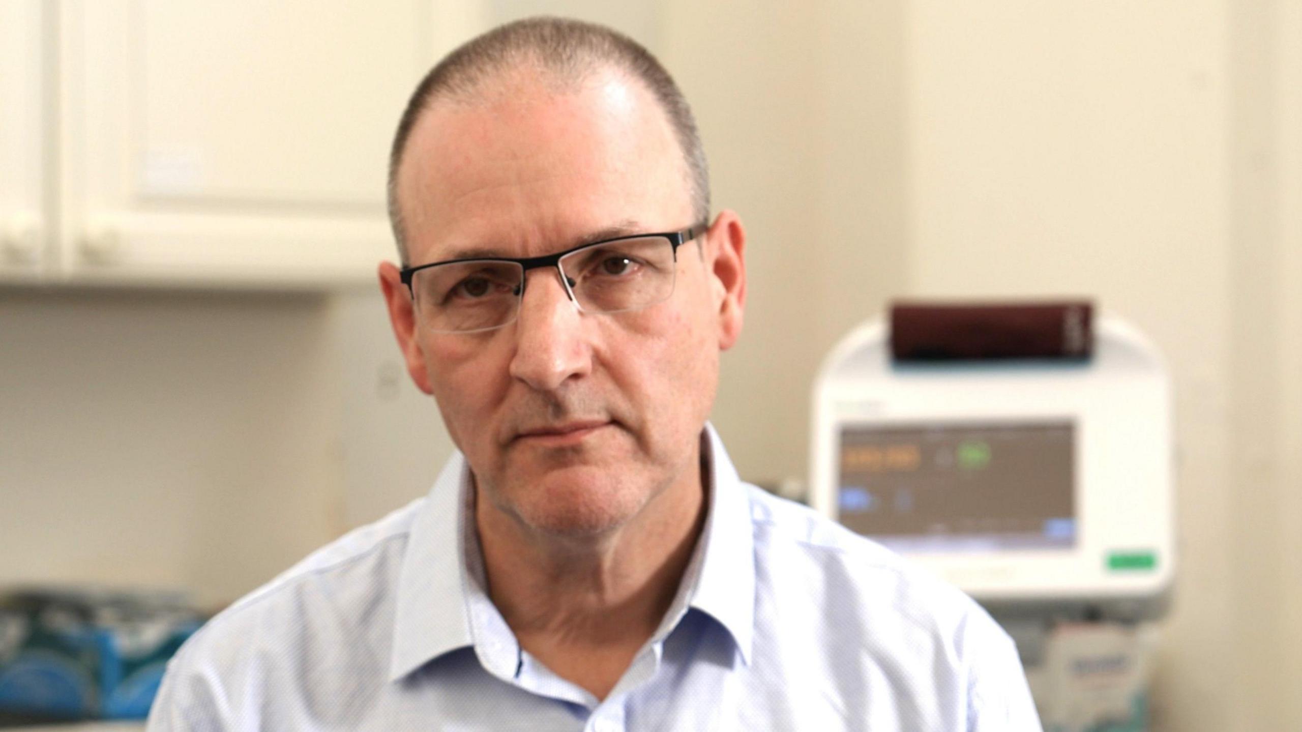 Martin Rolles looks at the camera. He wears a white shirt and glasses, and short dark hair. Medical equipment with a screen can be seen behind him.