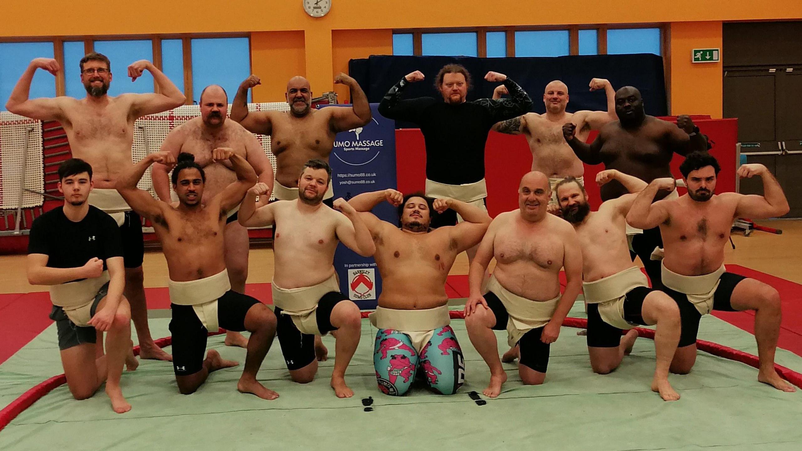 A group of men, all different ages, ethnicities and sizes. Most of them are topless and are flexing their muscles. They're all wearing a mawashi - the loincloth used while competing in sumo.