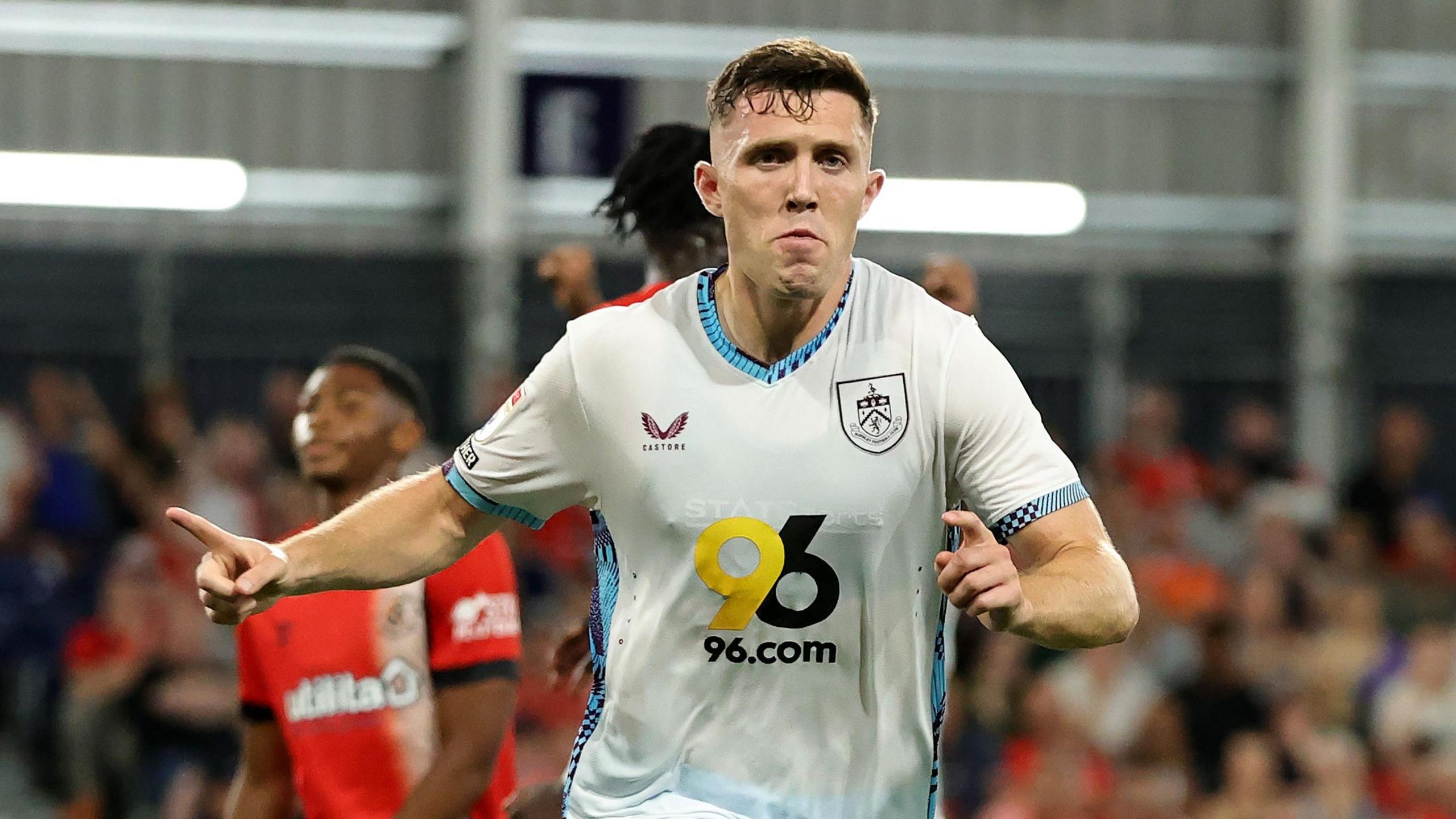 Dara O'Shea, wearing Burnley's white kit, holds his right arm out with his index finger pointed out as he runs away to celebrate scoring a goal at Luton