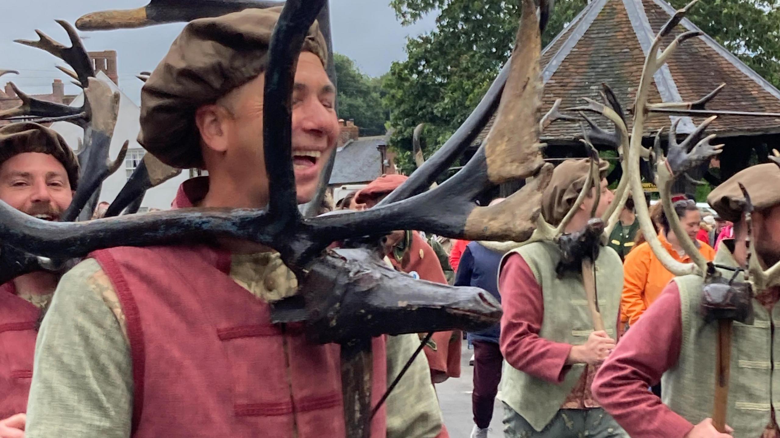 A close-up of the dancers holding the deer horns and carved wooden heads of the deers. They wear brown caps as well as light shirts and pink tunics or pink shirts with light tunics. The man closest to the camera is smiling broadly. 