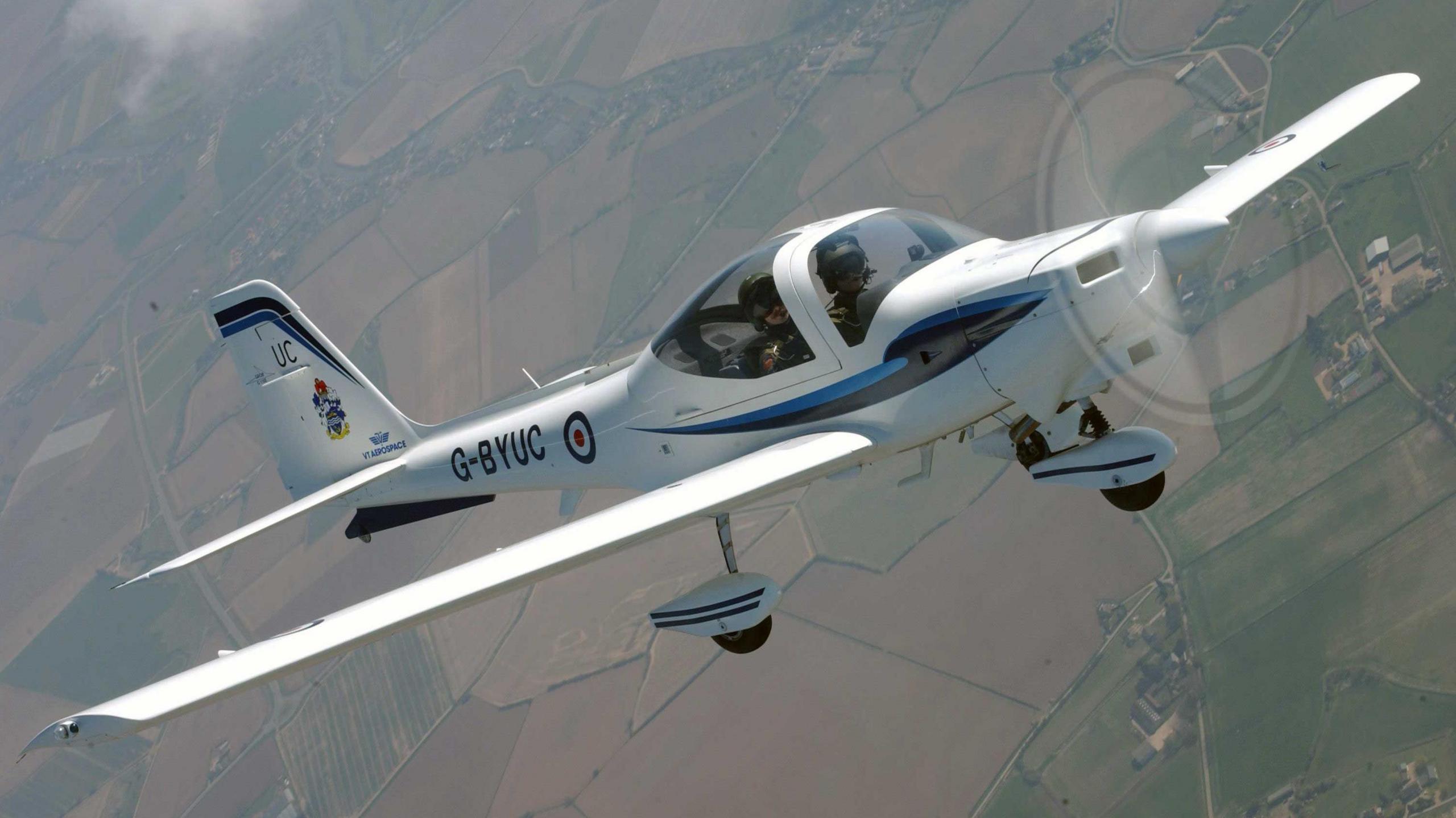 A small white RAF plane seating two people is seen mid-flight, with a view of fields below.