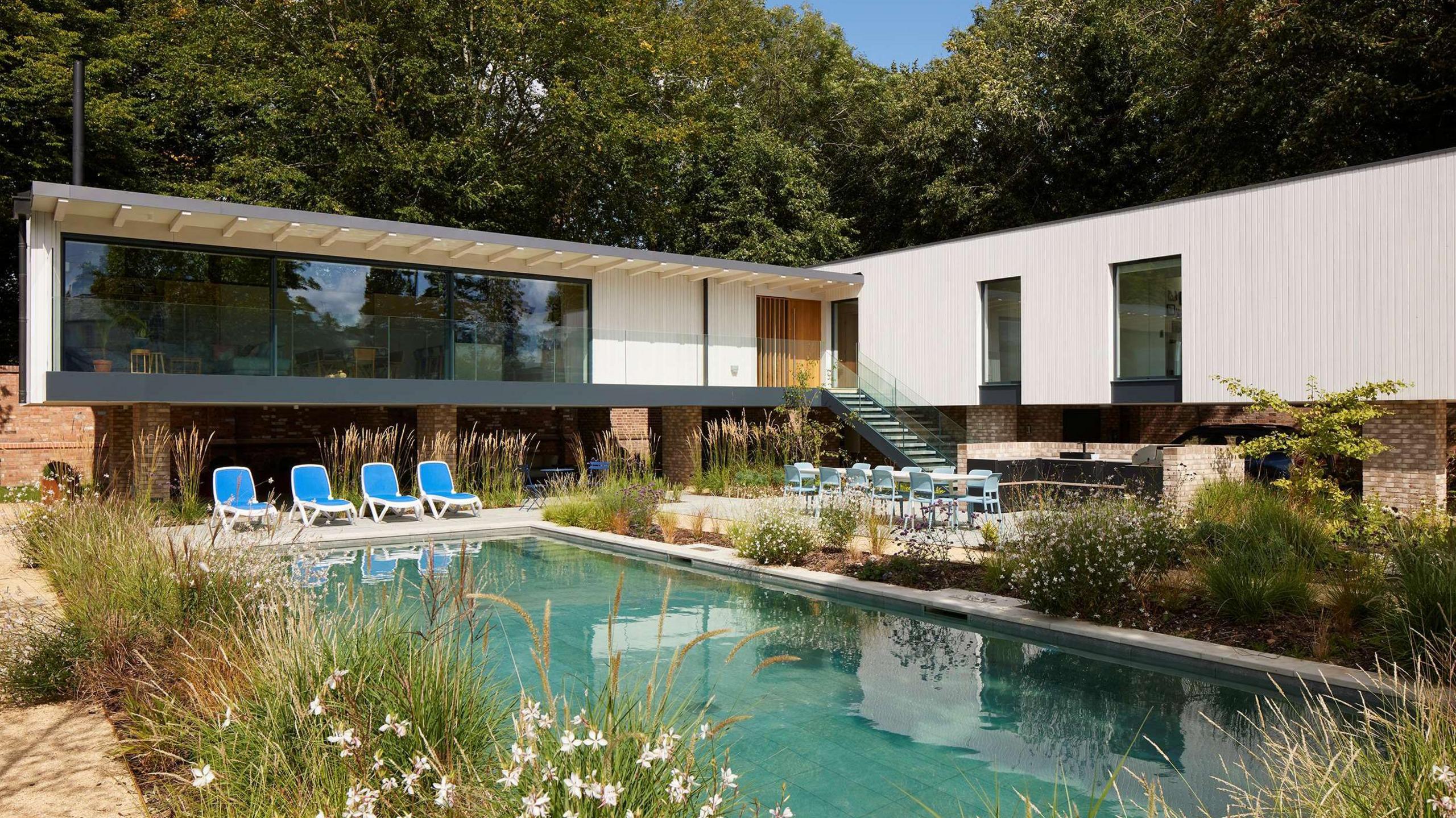 A modern white house that is one brick stilts, with a pool at the front. The pool is surrounded by plants and flowers and there are blue sun loungers in front of the house. 