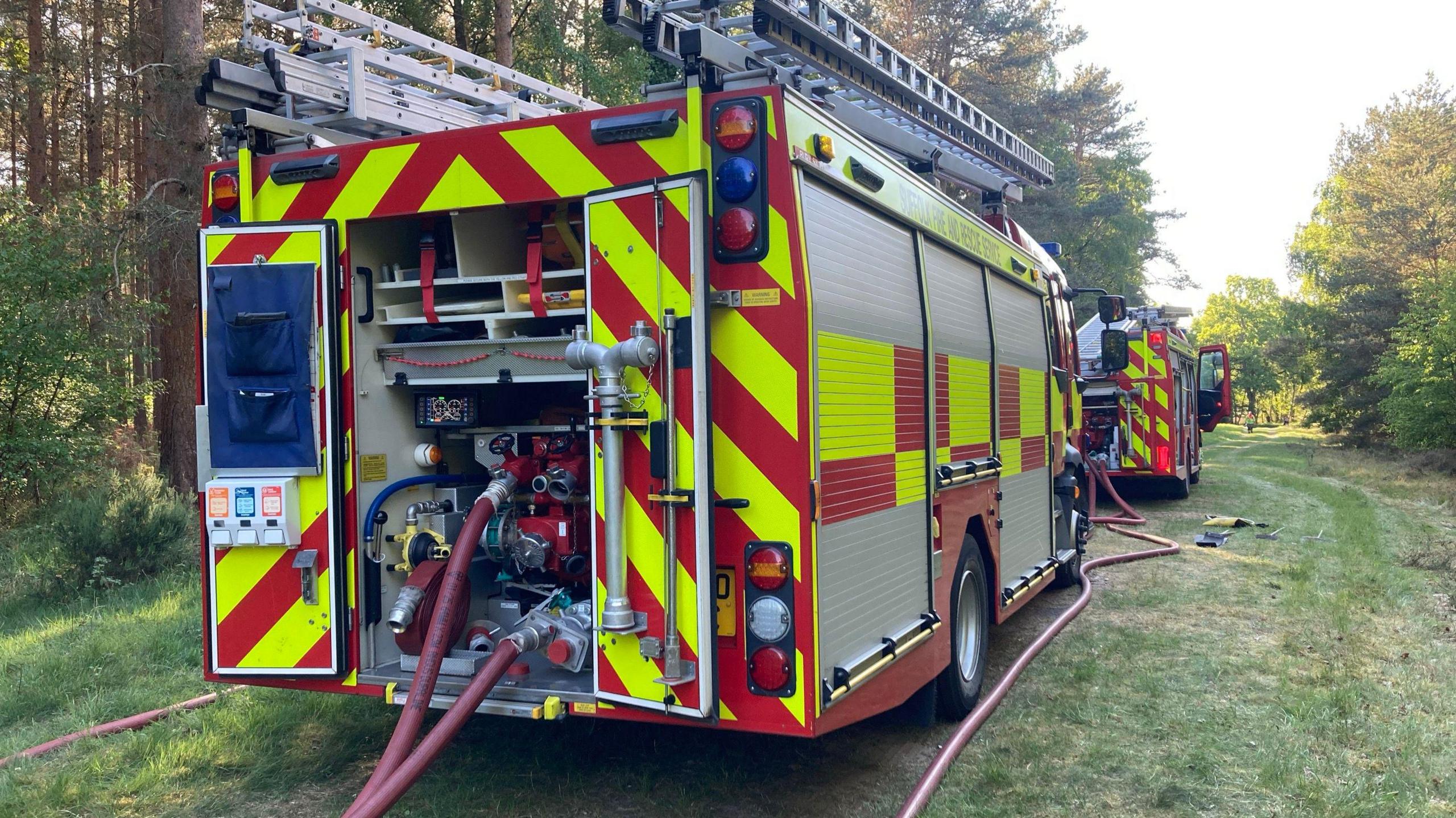 Two parked fire engines in a forest. The back doors are open on both and hoses have been attached. The red hose pipes are thick and have been lay along the grass .