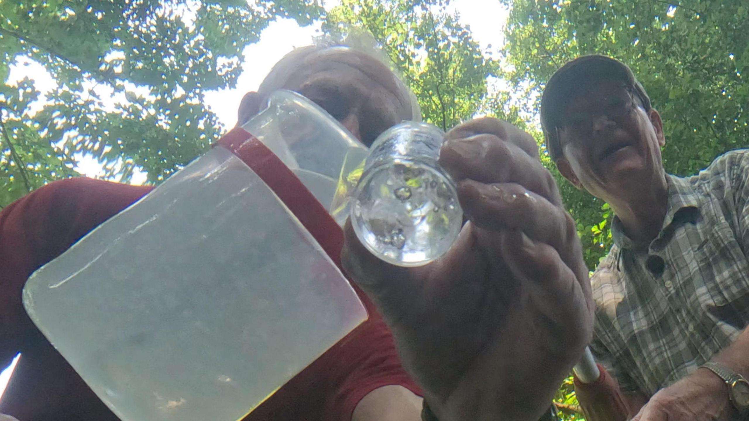 Two men shot from below one pouring river water into a glass jar with a leafy tree canopy behind them