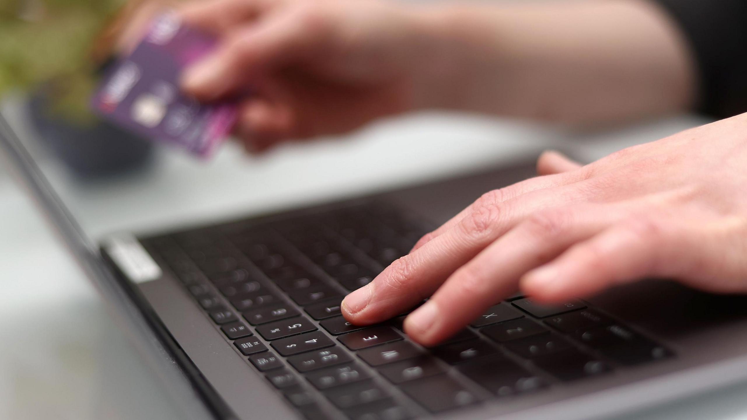 A woman's hands at the keyboard of a black laptop computer. Out of focus, she is holding a bank card in her right hand.