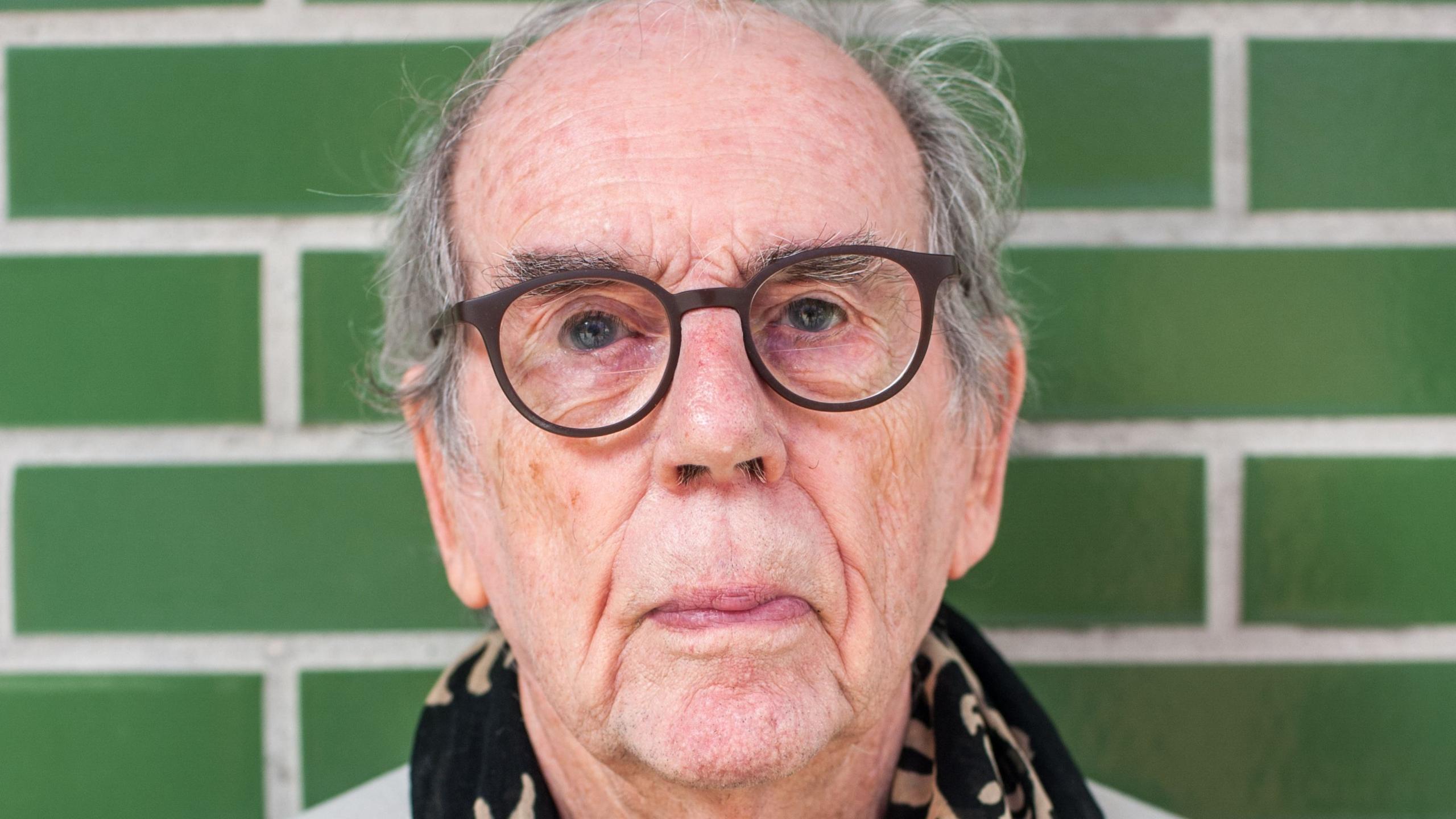 A man is looking at the camera. He is an older gentleman wearing black round-rimmed glasses. He has wispy grey hair and is stood in front of green bricks