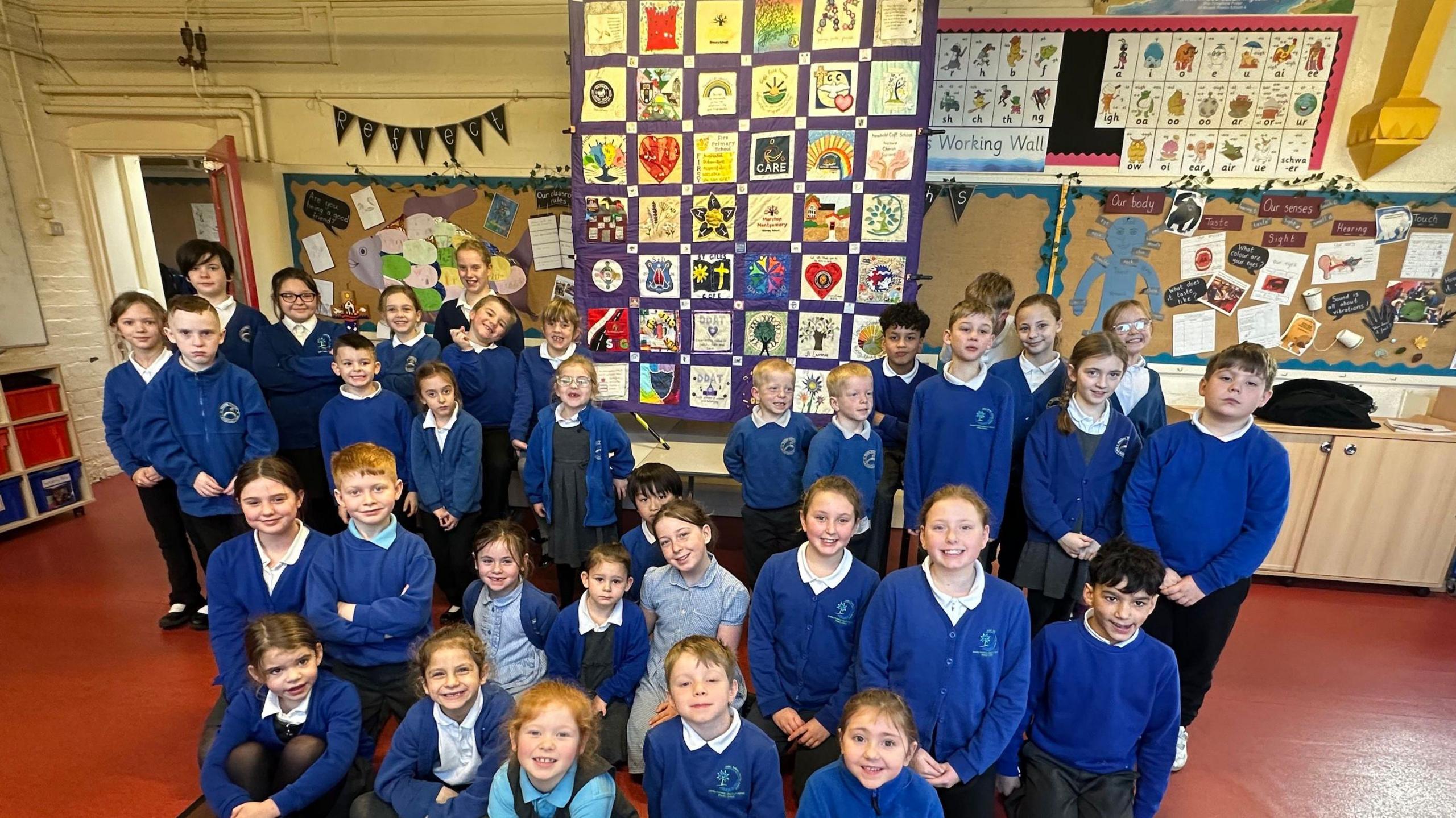 Multiple school children stand in a classroom in front of a quilt featuring patches with the logos of different schools.