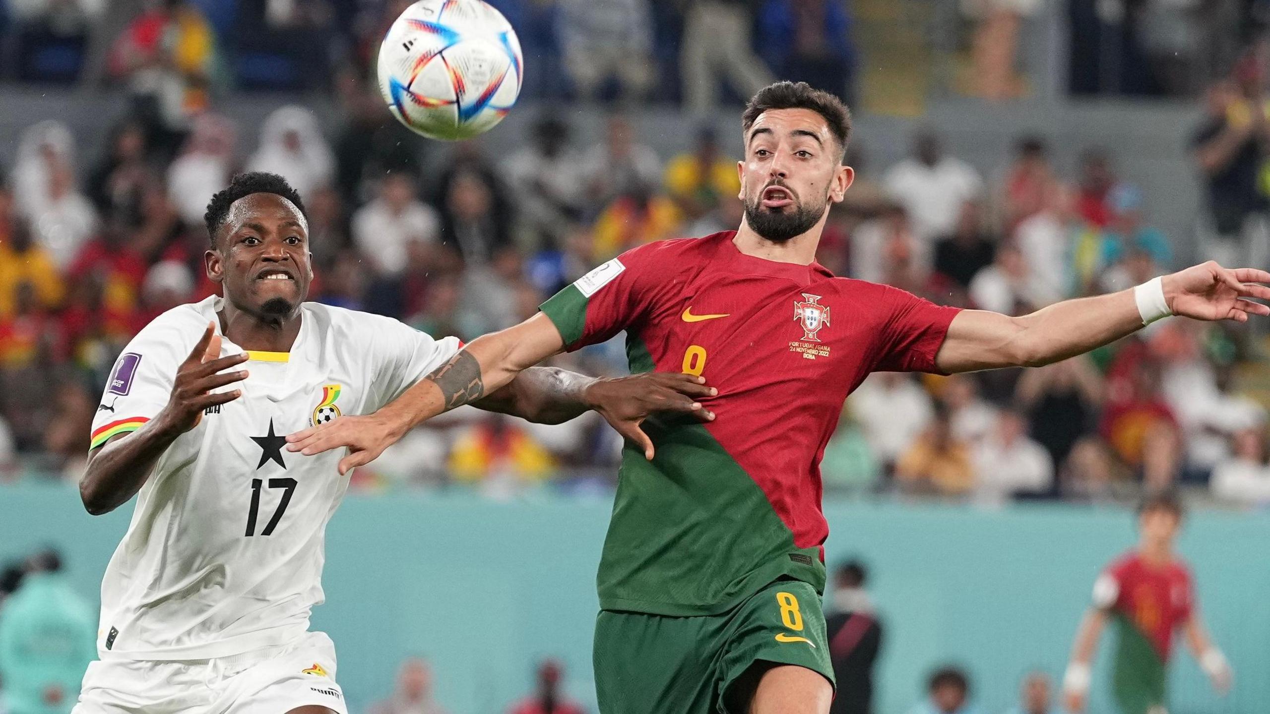 Baba Rahman, wearing a white Ghana kit, battles for a bouncing ball with Bruno Fernandes, who is wearing a red and green Portugal strip, during a game at the 2022 Fifa World Cup