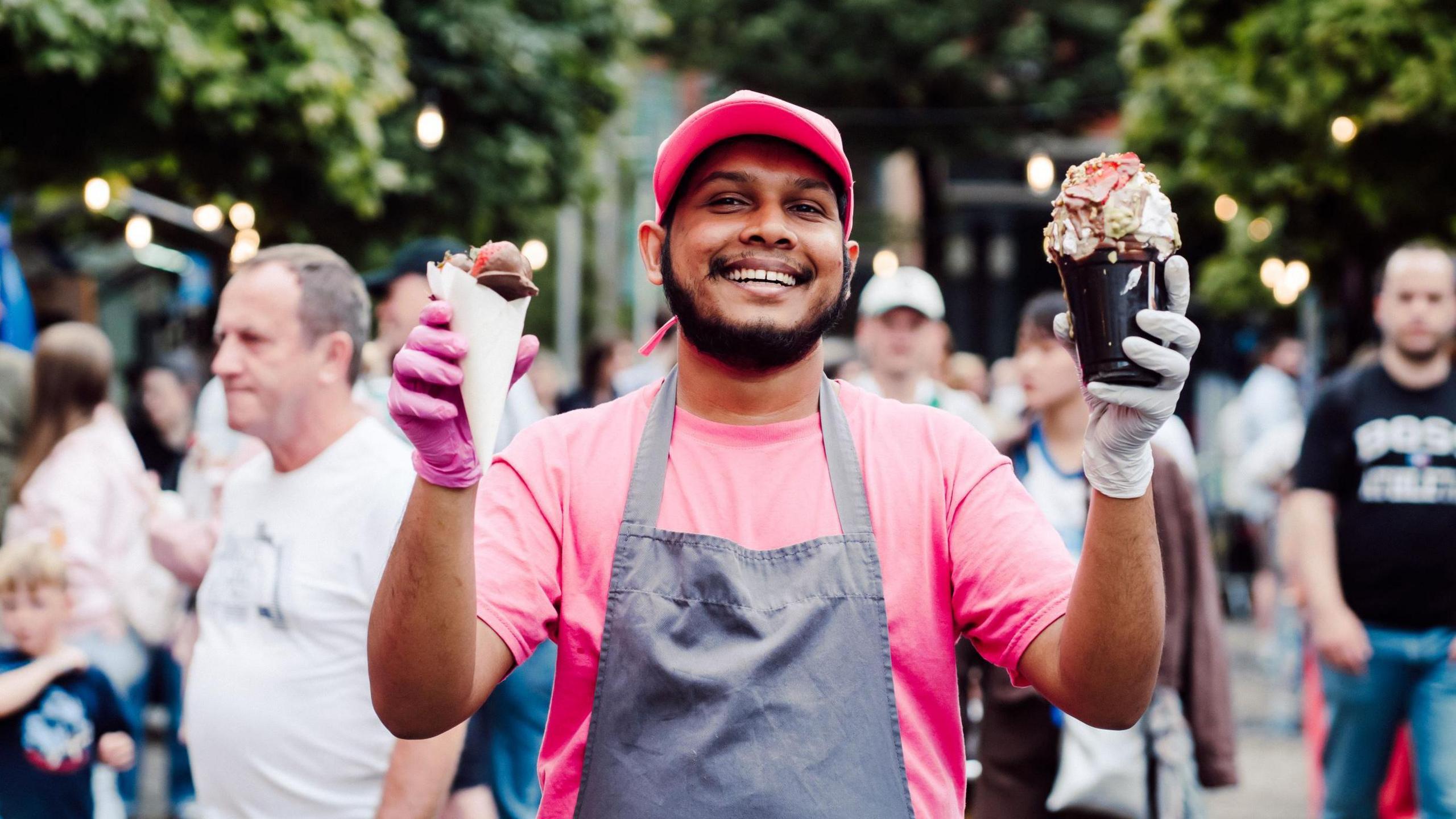 Man in pink t-shirt and hat wearing grey apron. He is holding icecream in both hands