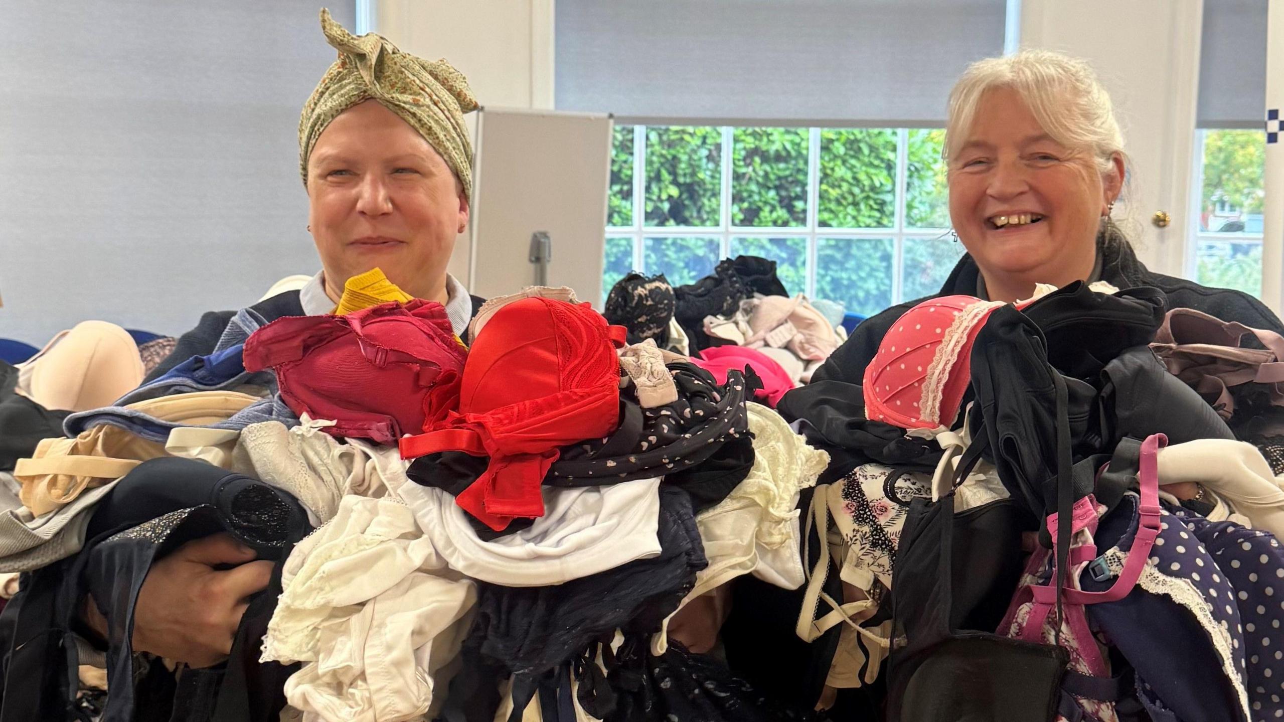 A woman in a headband and a woman with light grey hair smile and hold a huge pile of bras, ranging from bright red ones, to polka dot ones to silky ones.