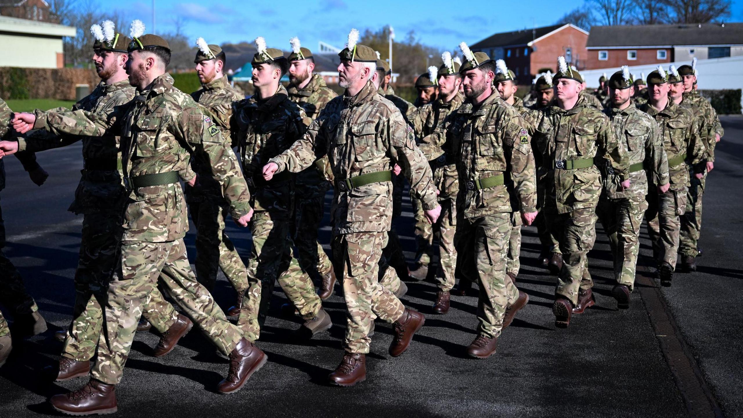 A group of male soldiers in berets and camouflage uniform march along a road. 