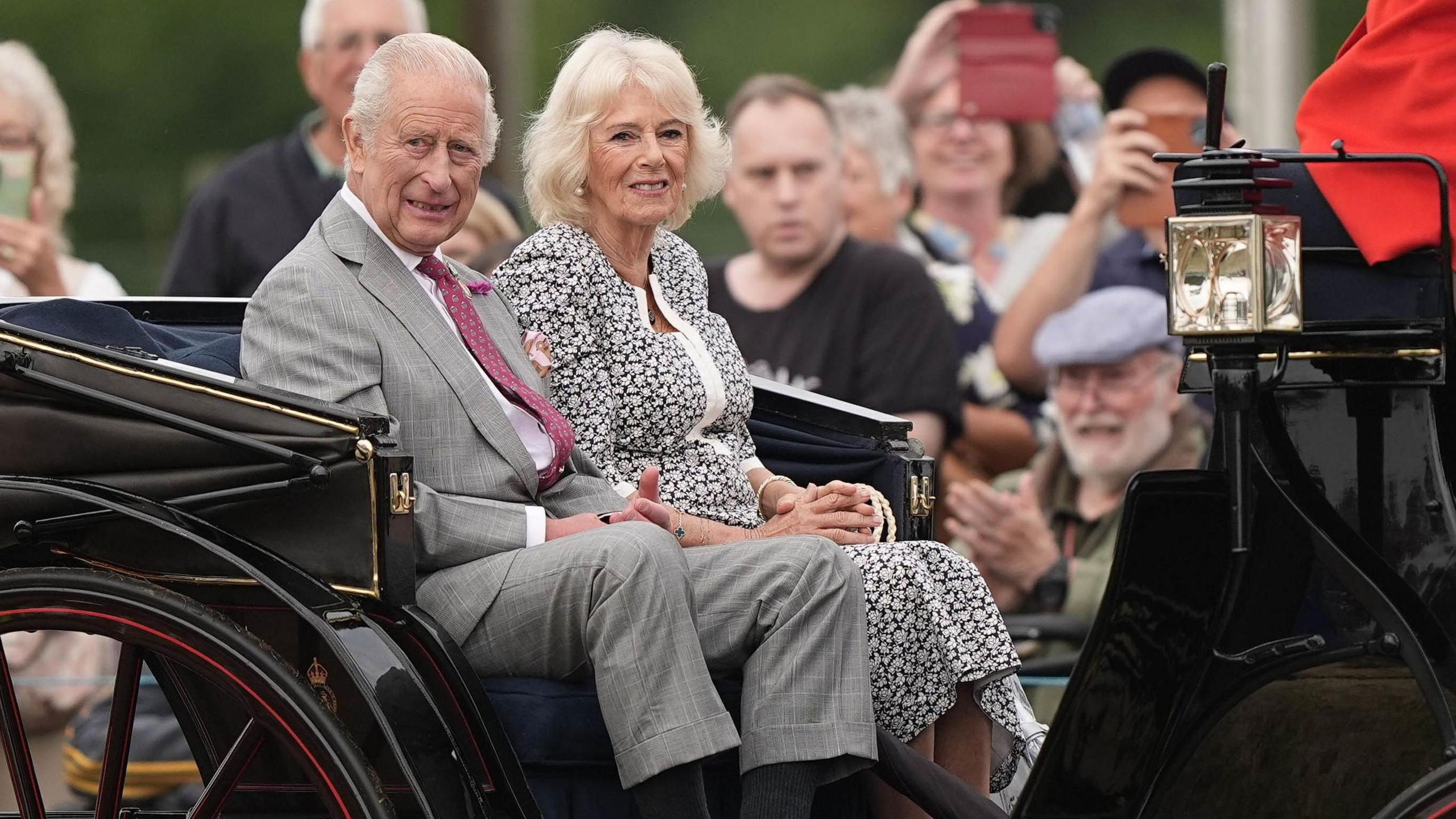 King Charles III and Queen Camilla during a visit to the Sandringham Flower Show at Sandringham House in Norfolk. They are in a black carriage being pulled by a horse. There are crowds in the background.