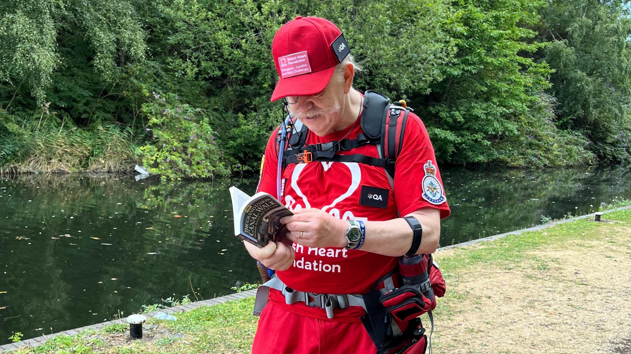 David stands in front of a canal. He is wearing a rucksack and reading a book. The title of the book is "The Grand Union Canal Walk"