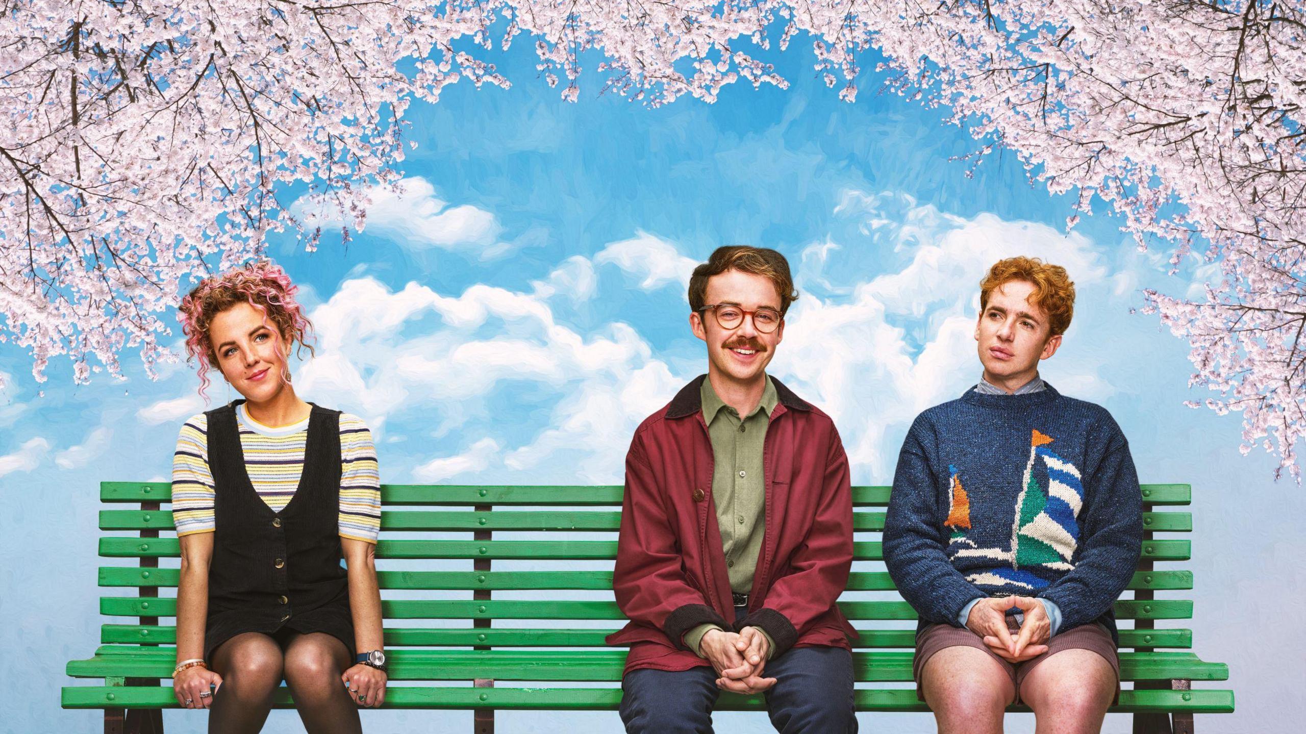 A woman with long pink curly hair sits to the far left of a green bench. She wears a black yellow and blue stripy t-shirt under a black buttoned dress. Behind her is a partially cloudy blue sky with pink blossom trees either side of the bench, hanging over it. To the right of the bench are two men. The man in the middle has short brown hair with red clear spectacles, he wears a green shirt, red jacket and navy jeans. The man on the far right wears a navy sweater with an illustration of a yacht on it. the yacht has a green white and blue sail. The man also wears a pair of brown shorts.