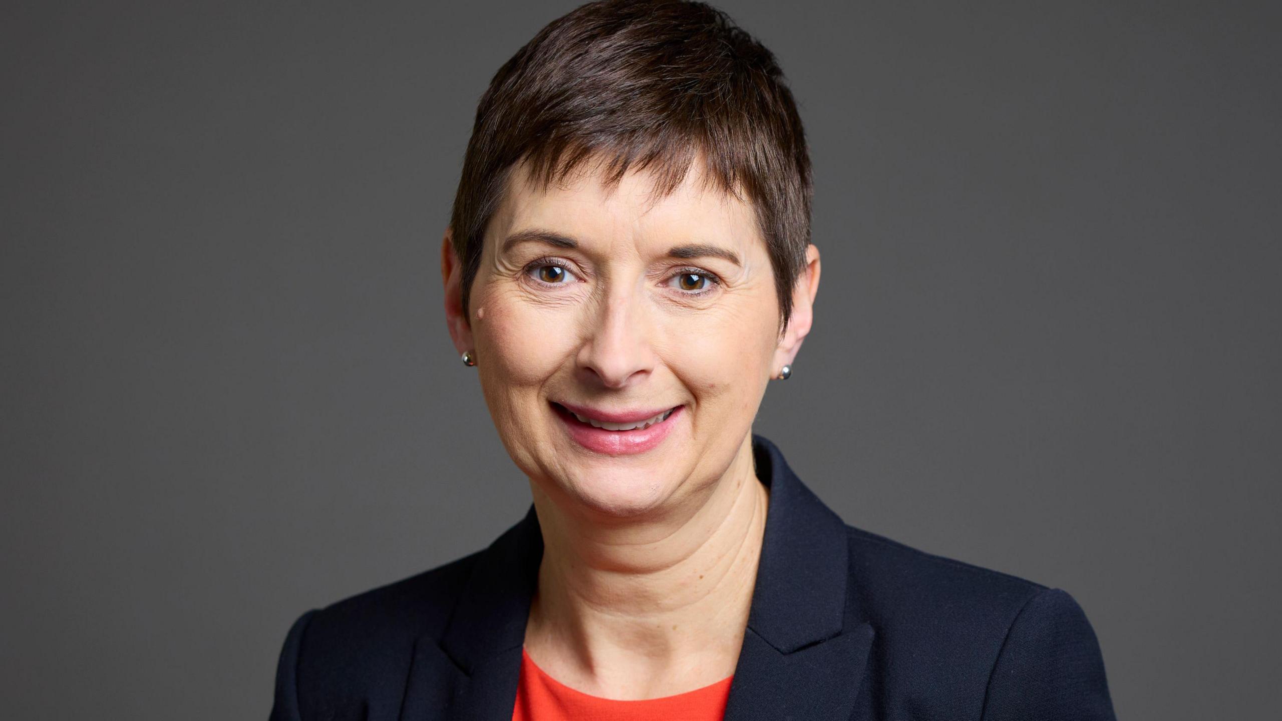 A woman with short dark hair, a black jacket and red top, smiles while standing in front of a dark grey background.