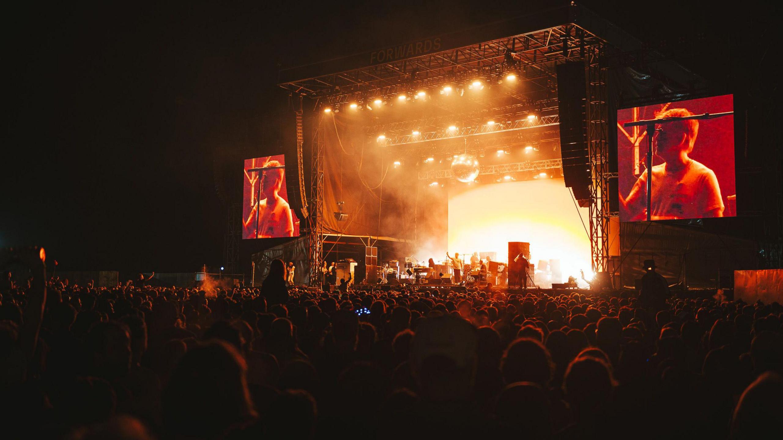 A dance group perform on stage at Forwards Festival in Bristol on the Downs. The large crowd is illuminated by orange floodlights and the performers are visible on large screens either side of the stage