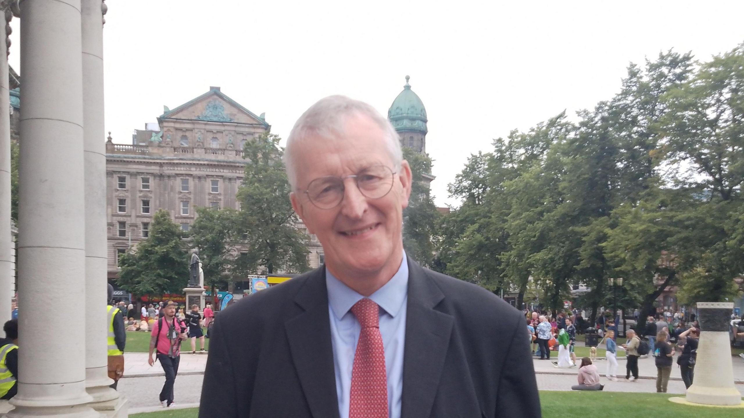 Hilary Benn MP standing outside Belfast City Hall, wearing a suit and red tie. He has grey hair and glasses