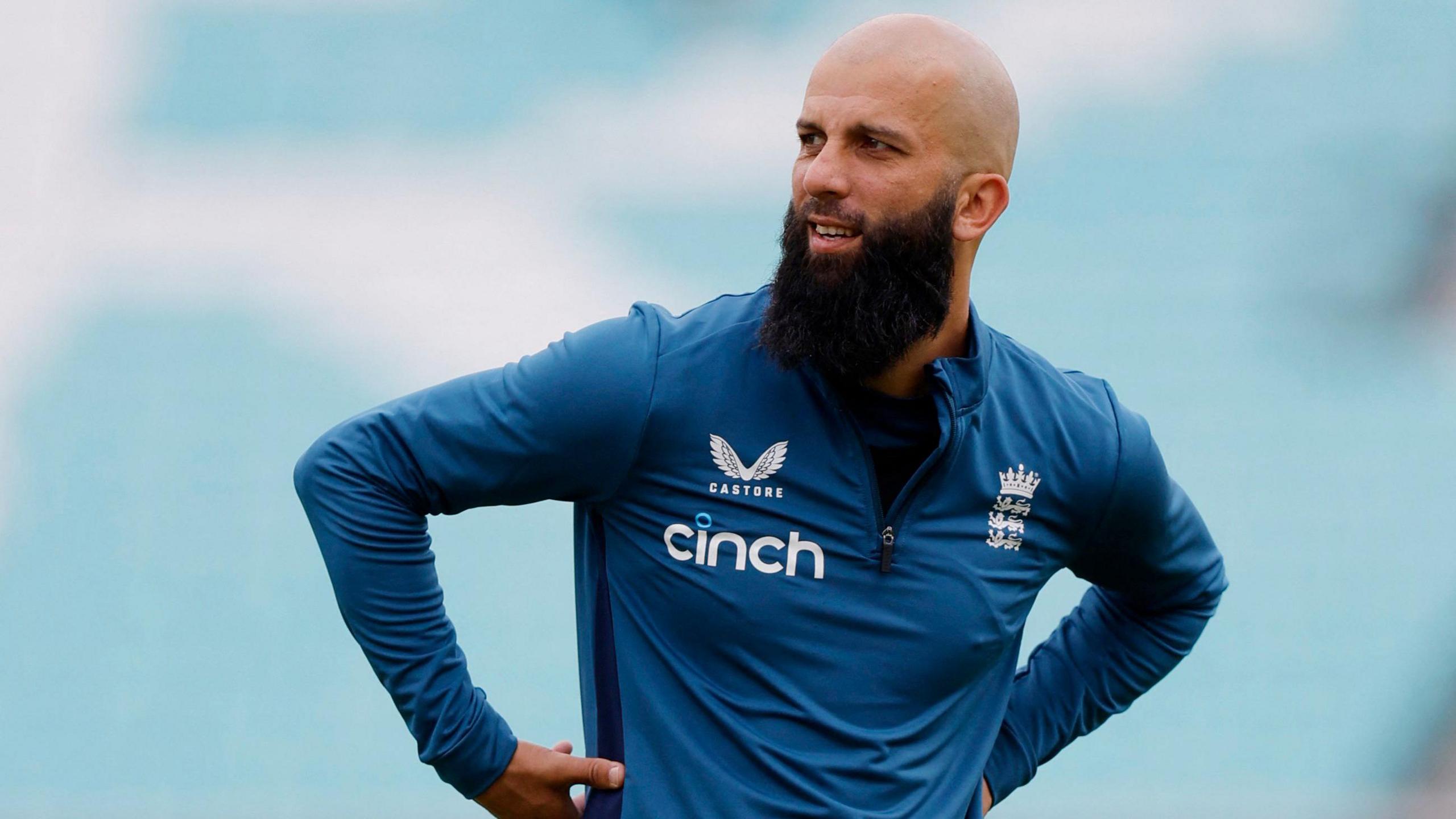 Moeen Ali has a large black beard and is bald. He is wearing a blue England cricket long sleeved top