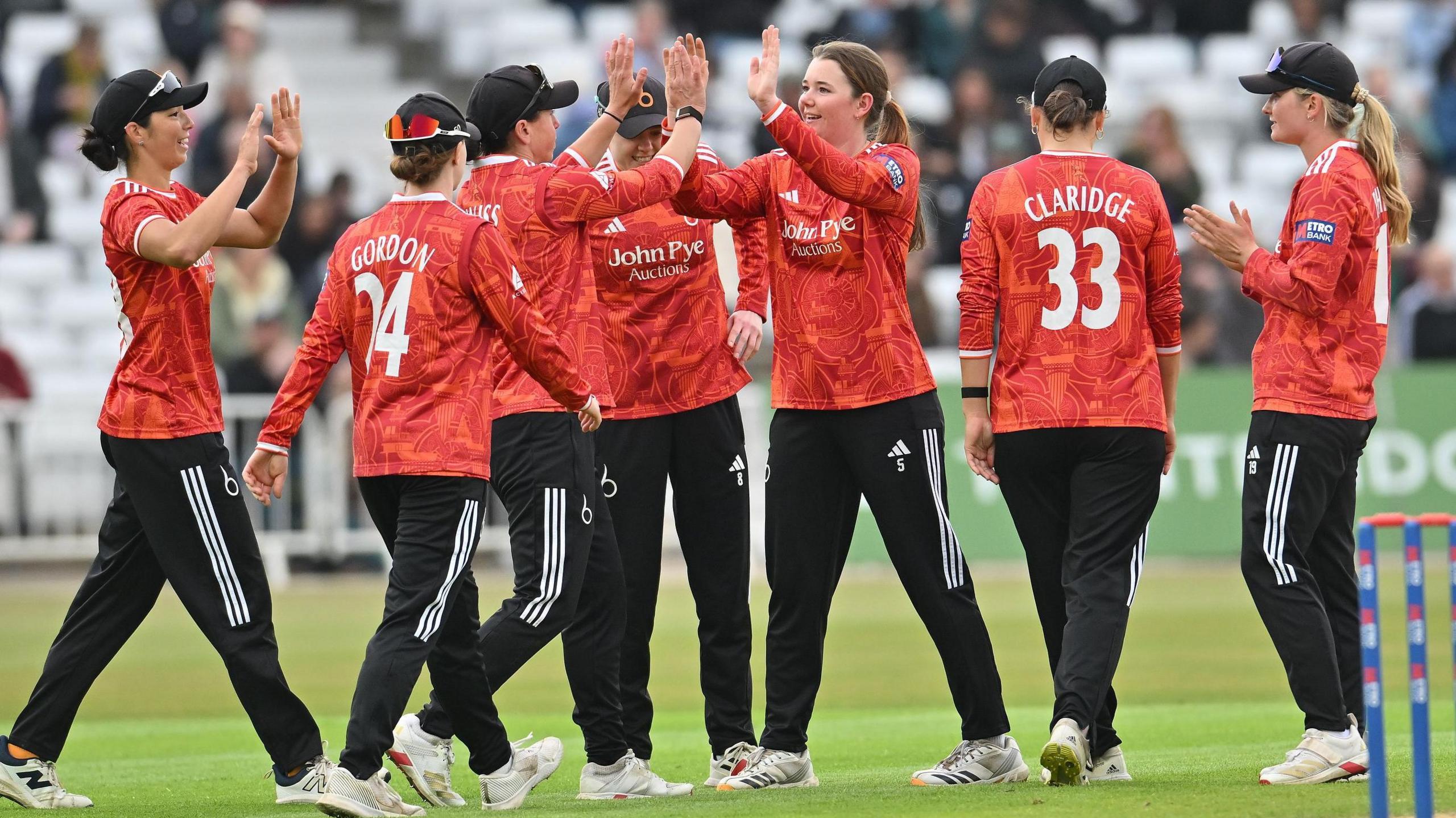 Seven team-mates from The Blaze celebrate a wicket from Josie Groves at Trent Bridge. Josie Groves is central and is high-fiving Georgia Elwiss, with the other team-mates around them. They are wearing the Blaze 2026 Orange One-Day Cup kit with black trousers.