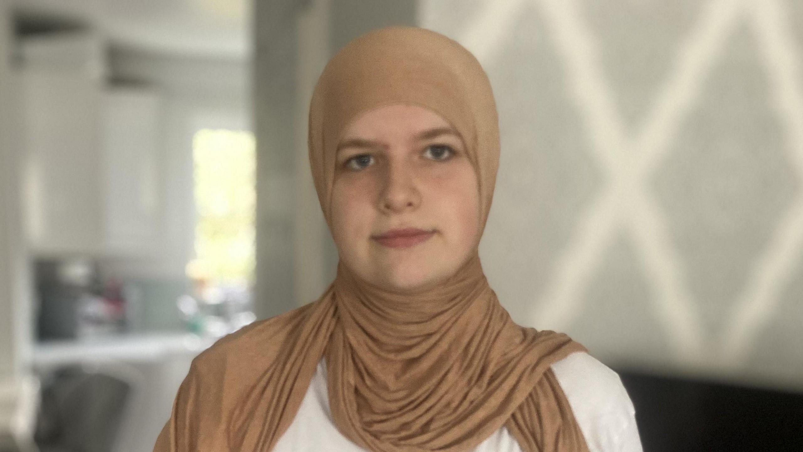 Aleyah Knight. She is pictured in a house wearing a beige hijab and a white t-shirt. She is looking at the camera and smiling