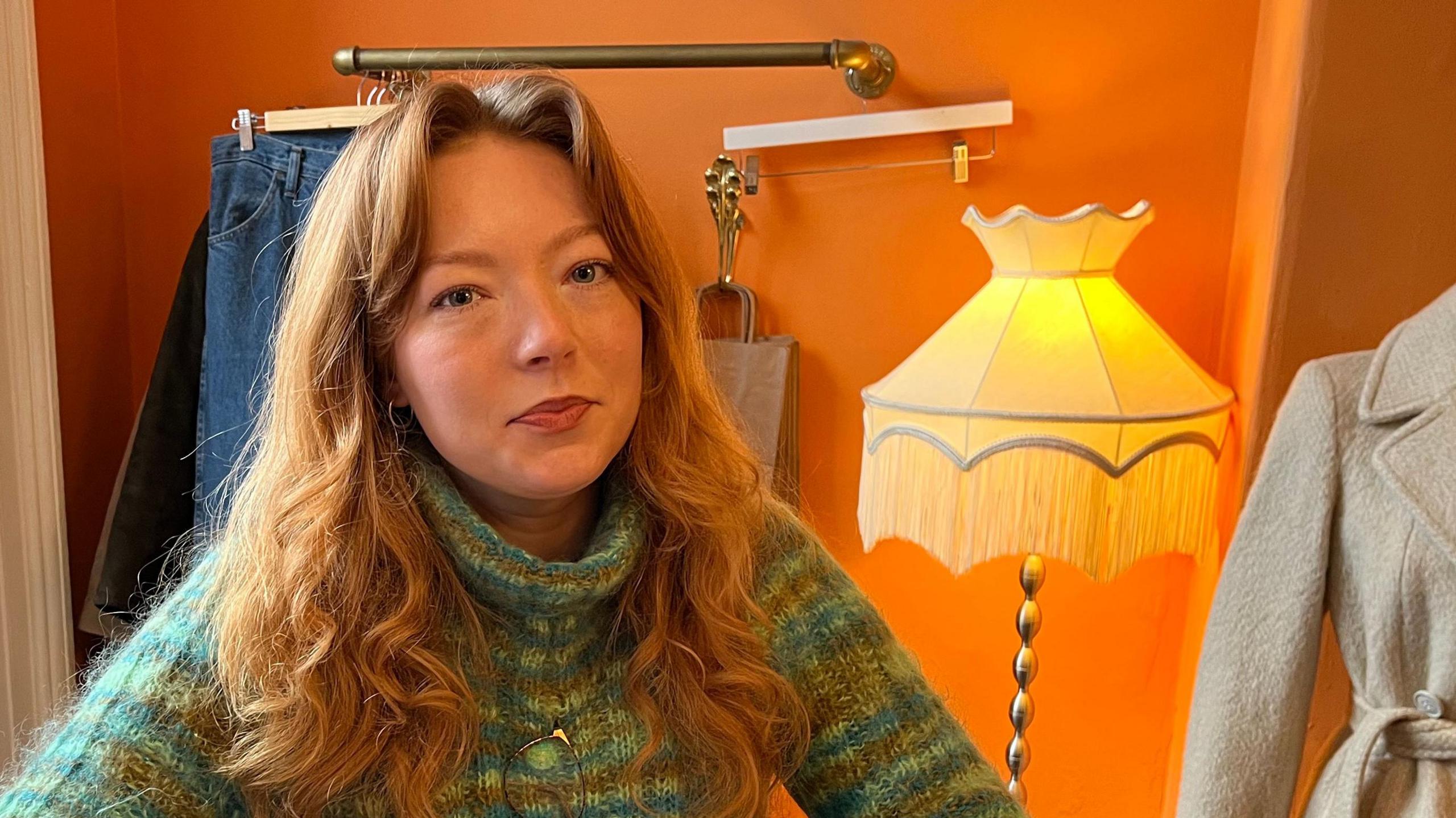 Laura Jackson. She is standing behind the counter of her shop and has long red hair and is wearing a green knitted jumper. A stylish coat and an ornate lamp are displayed behind her.