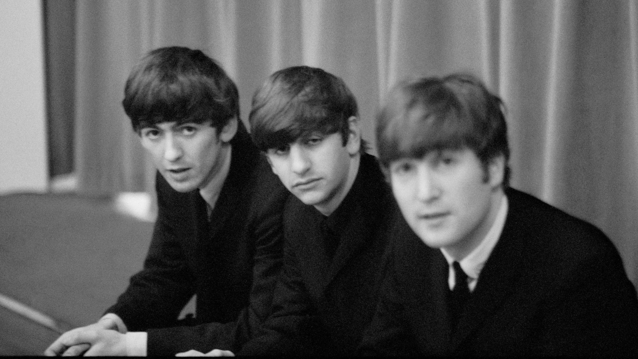 Titled: George, Ringo, and John backstage at The Beatles Christmas Show, Finsbury Park Astoria, London, December 1963. The three bandmates are sat down and look down the lens of the camera. They wear dark suits and are captured in black and white film.