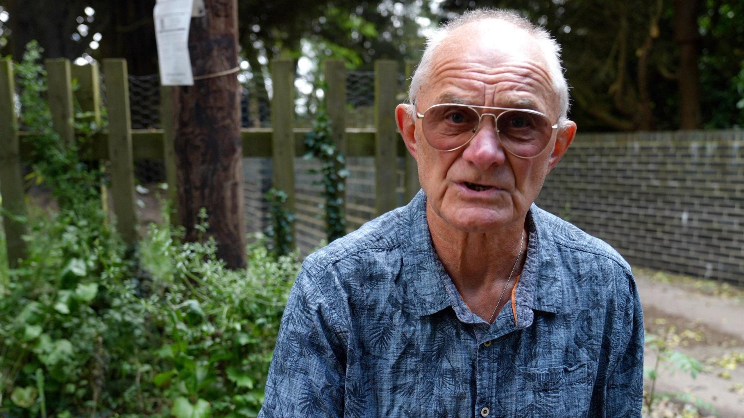 A man with short white hear wearing a blue shirt and tinted glasses. He is kneeling by a water leak in a country lane.