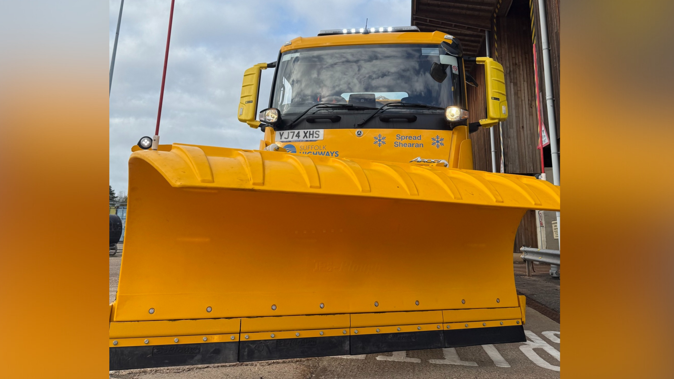 A close-up of the front of a bright yellow gritter lorry, which shows its cab and snow plough. 
