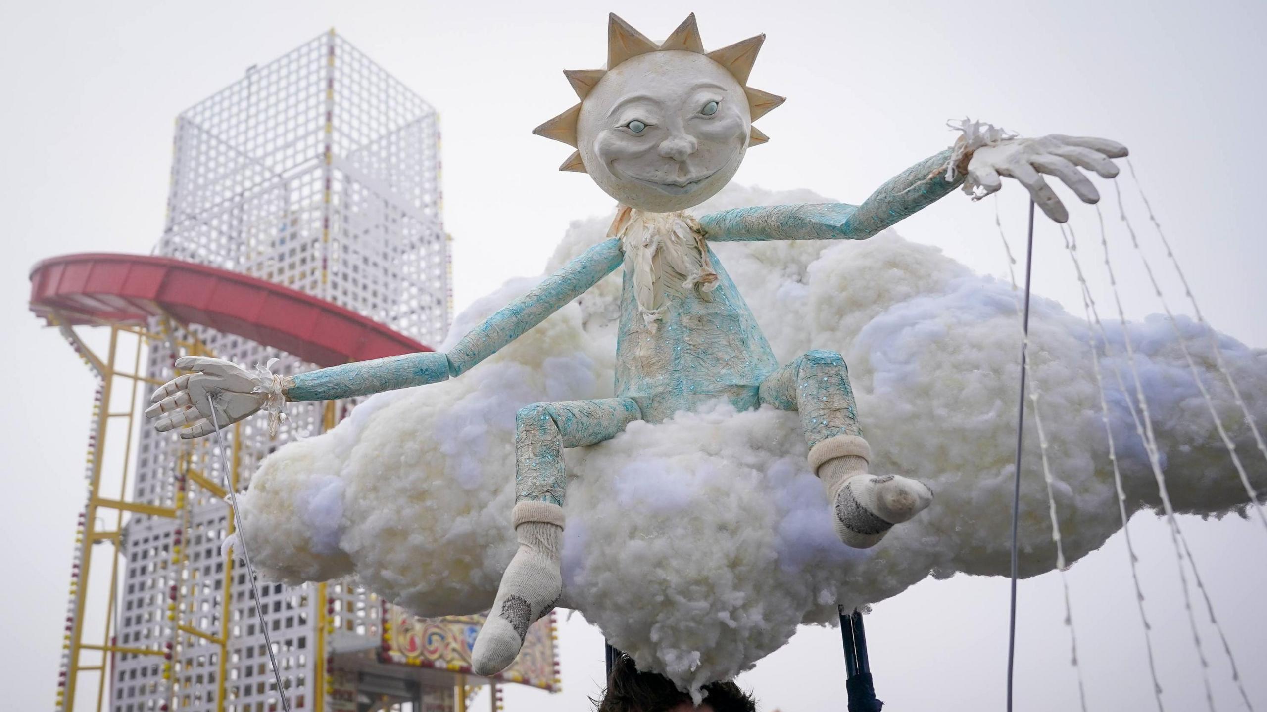 A street art puppet of a strange sun headed figure in blue sat on a cloud. A helter skelter can be seen in the background.