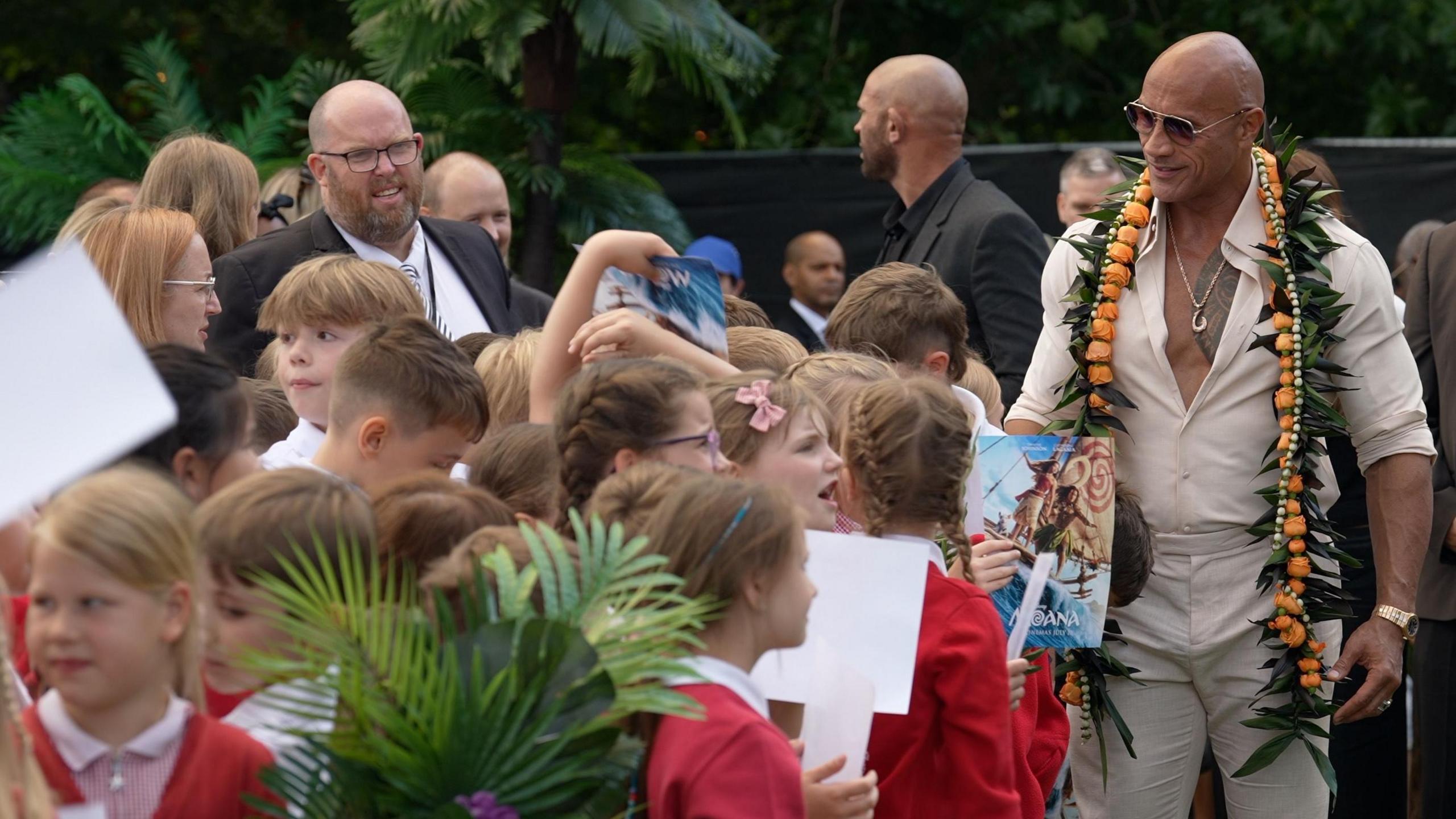 A radical  of schoolchildren wearing reddish  uniforms lasting  successful  beforehand   of The Rock – a antheral   wearing a pick  jumper and achromatic  trousers and sunglasses.