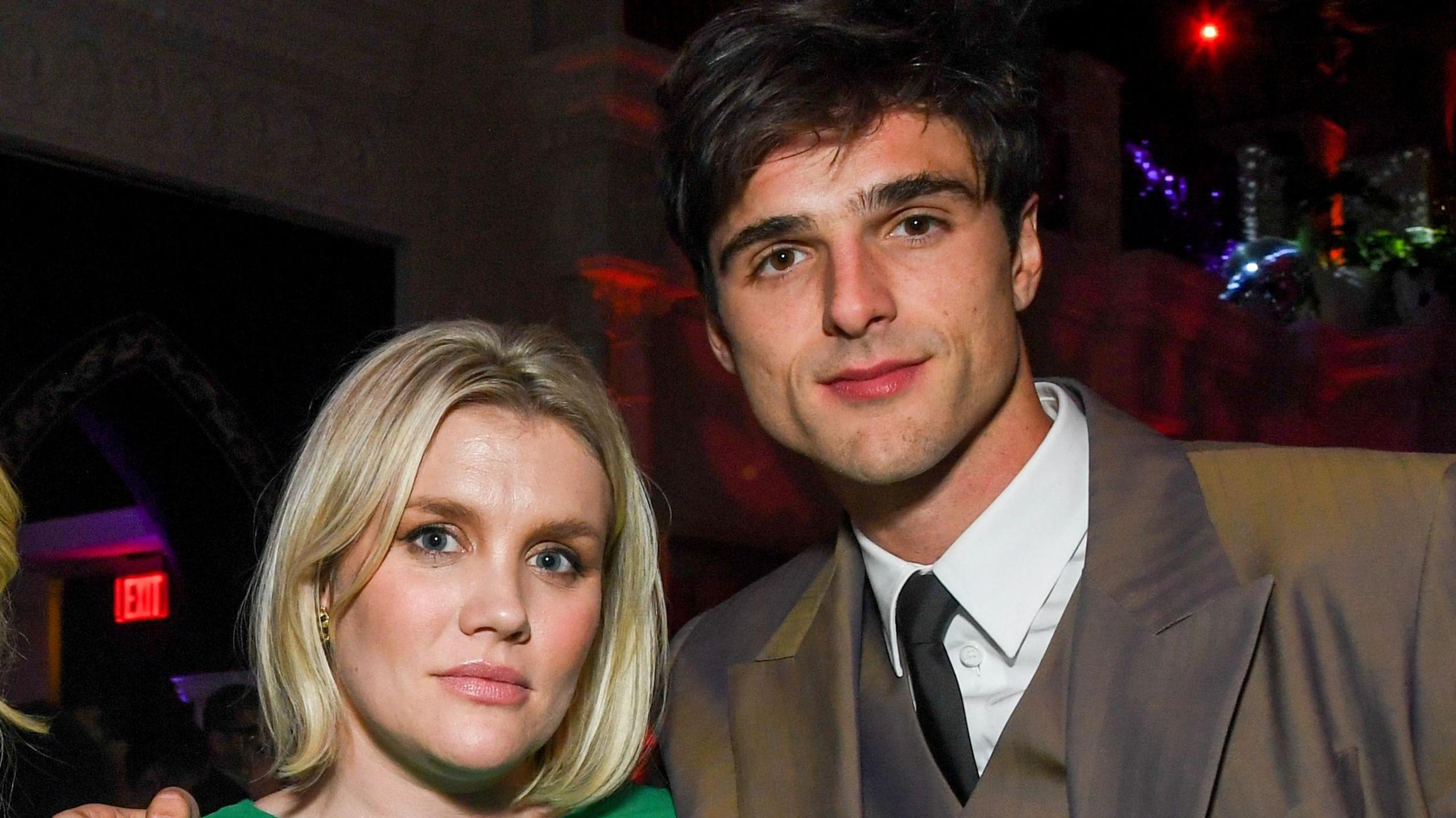 Director Emerald Fennell pictured with Jacob Elordi. Fennell has a chin-length blonde bob and is a woman aged about 40. Elordi, who has dark hair and eyes, has his arm around her shoulders as they both face forward posing for the camera. He is wearing a three-piece grey suit and a black tie.