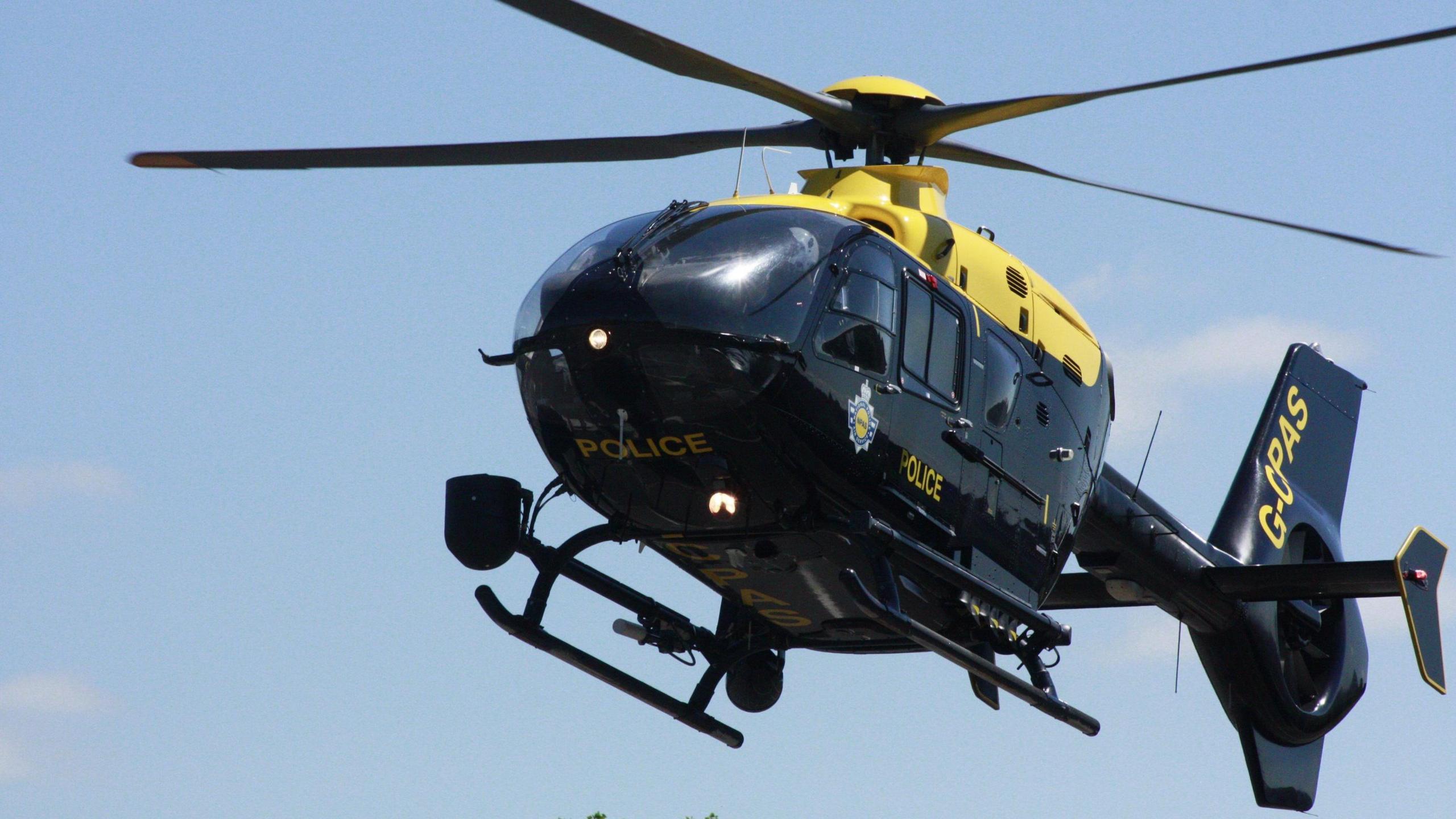 Exeter's NPAS helicopter flying in a blue sky. It is mostly back with a yellow strip at the top and it features a police shield on the side, the word police in yellow on the underside of the nose and the door and its aircraft number on the tail