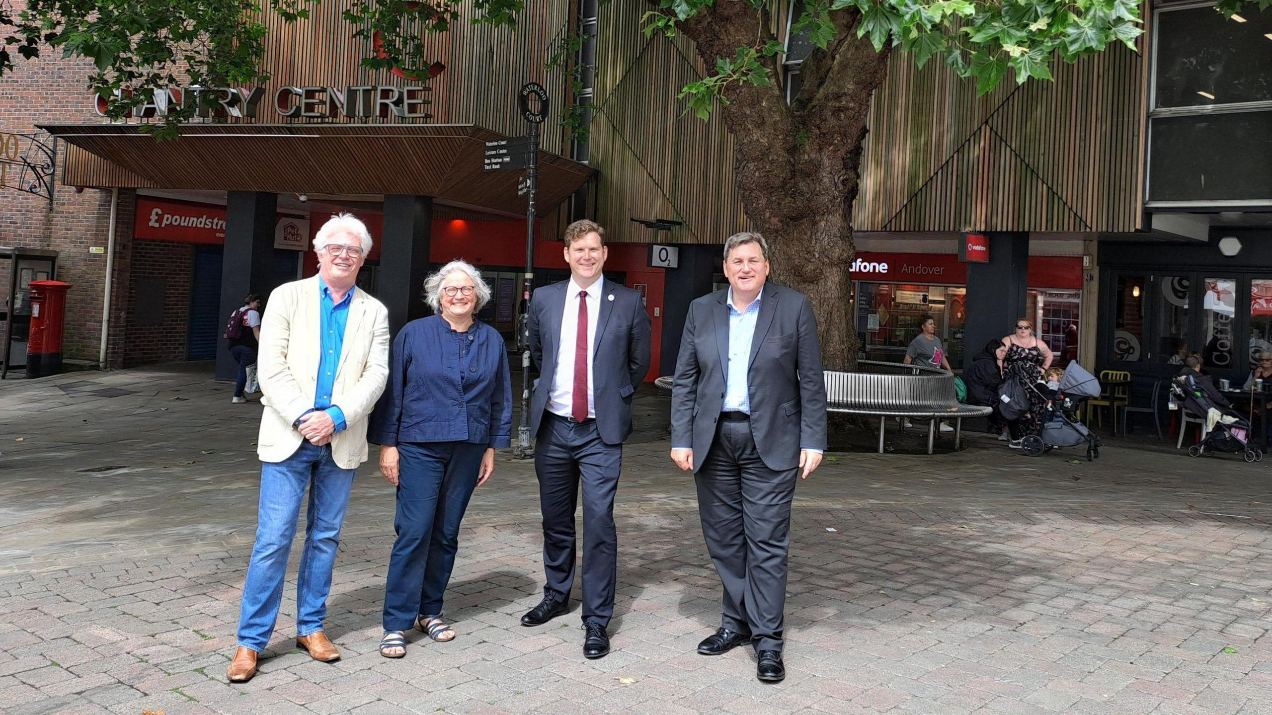 From left to right Aidan Ridyard and Helen Grassly of BFF, councillor Phil North and councillor Kit Malthouse MP, outside of the location for the new theatre in Andover. They are smiling at the camera. The Chantry Centre is in the background. A woman with a pram appears to be speaking to another woman who is sitting on a round metal bench. A man is passing by a shop.   