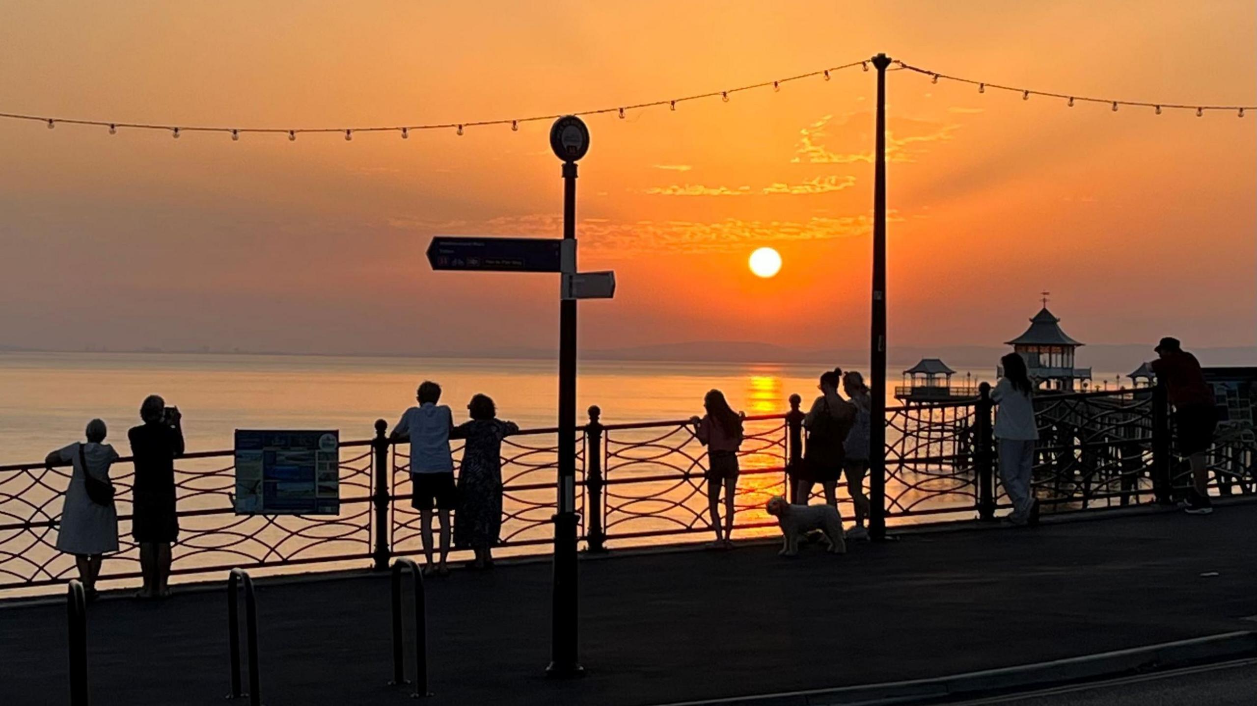A small group of people stand on the seafront at Clevedon at sunset. The sky is a dramatic mixture of oranges, with the sun glowing white in the sky. The people are silhouetted black against the sky and sea, as is part of the town pier