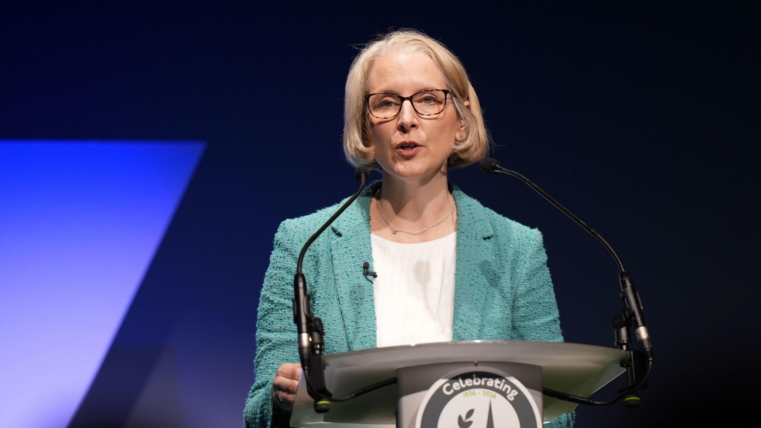 Reynolds stands at a podium, addressing the conference, in a green jacket and lit up in front of a purple background