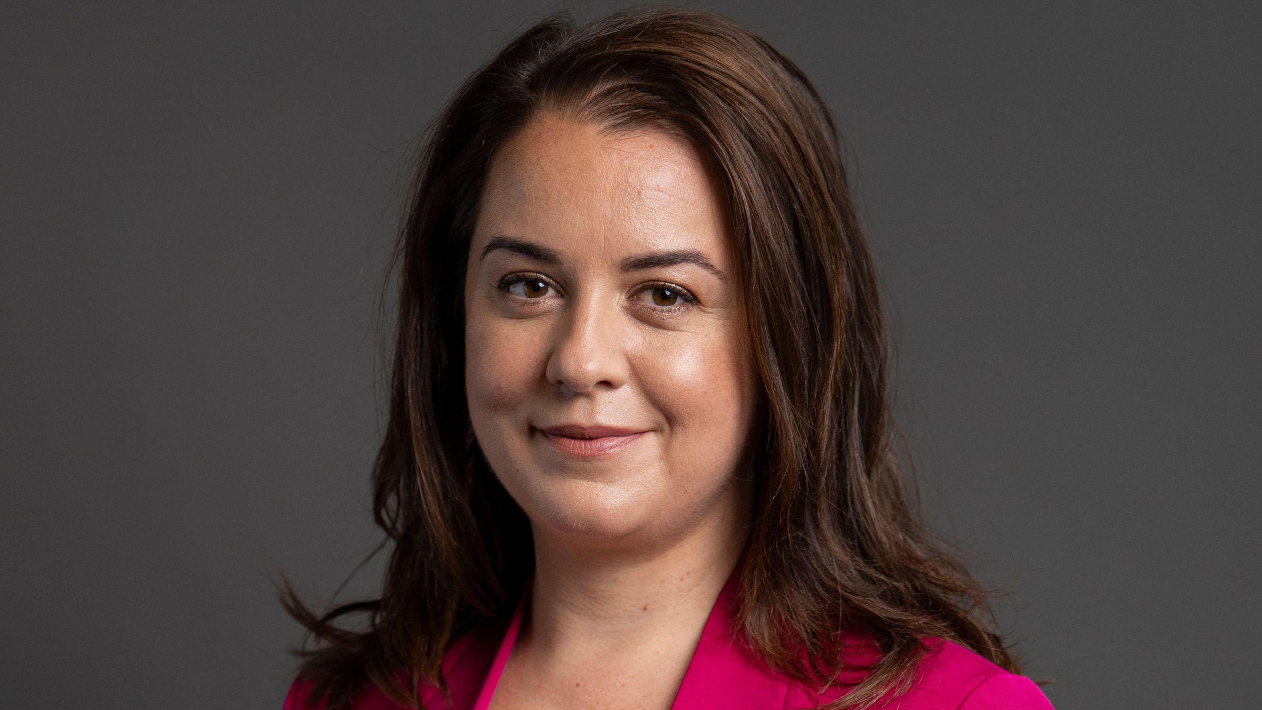 Parliamentary portrait of Culture Minister, Stephanie Peacock. She has shoulder length brown hair, and is wearing a pink jacket. Standing in front of a blank grey background.