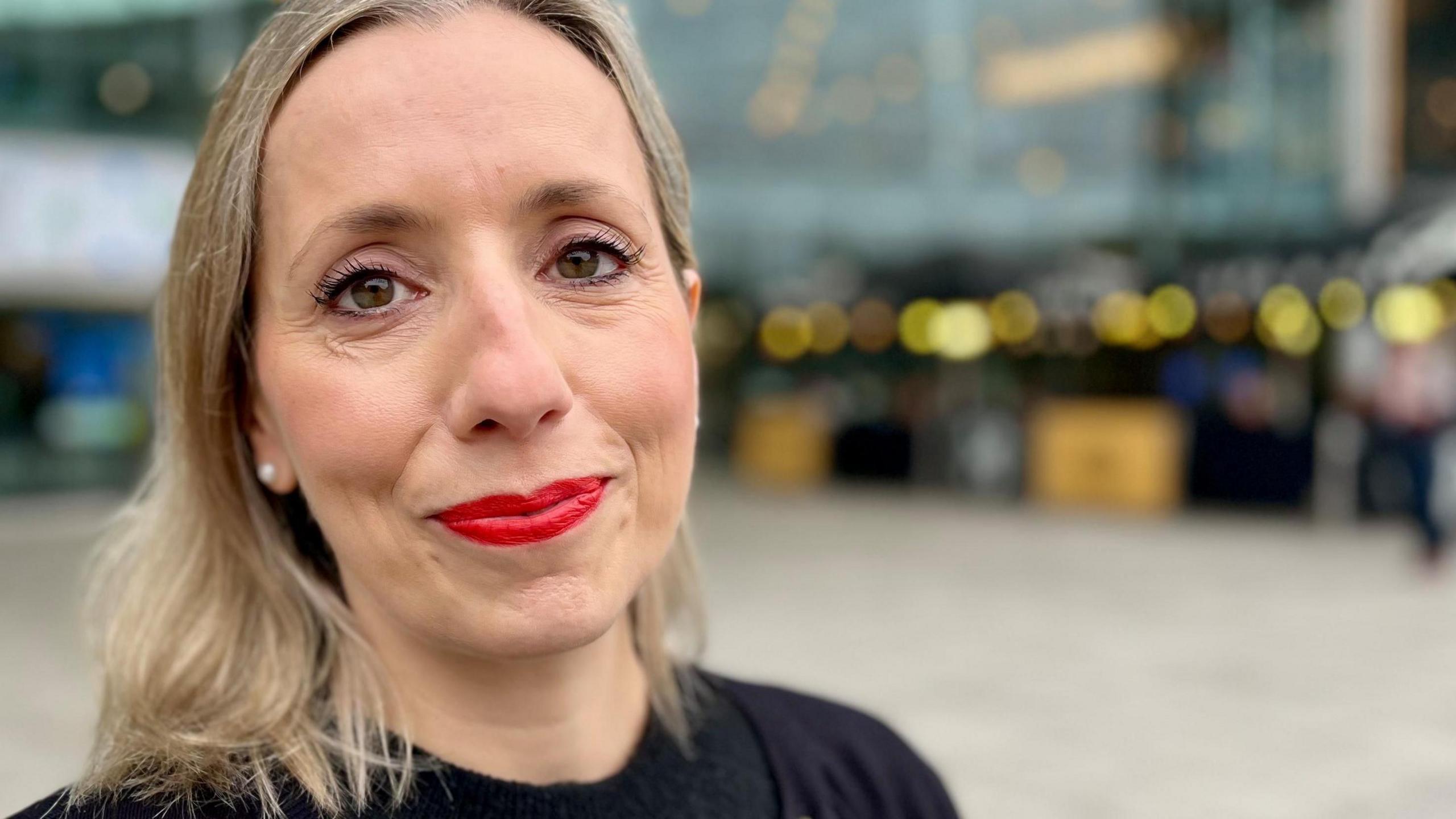 Sarah Taylor ka flokë bjonde deri në supe dhe mban veshur një bluzë të zezë dhe një xhaketë të gjatë. Ajo po buzëqesh në kamera dhe është fotografuar jashtë një ndërtese me panele xhami.