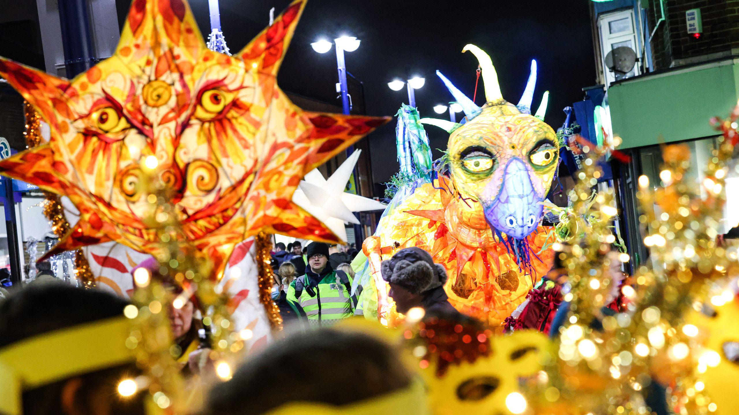 A previous year's illumination parade in Redcar. Two large illuminated puppets are being carried through the street. One has a large round face with spikes coming out of its head. The other is a large dragon.