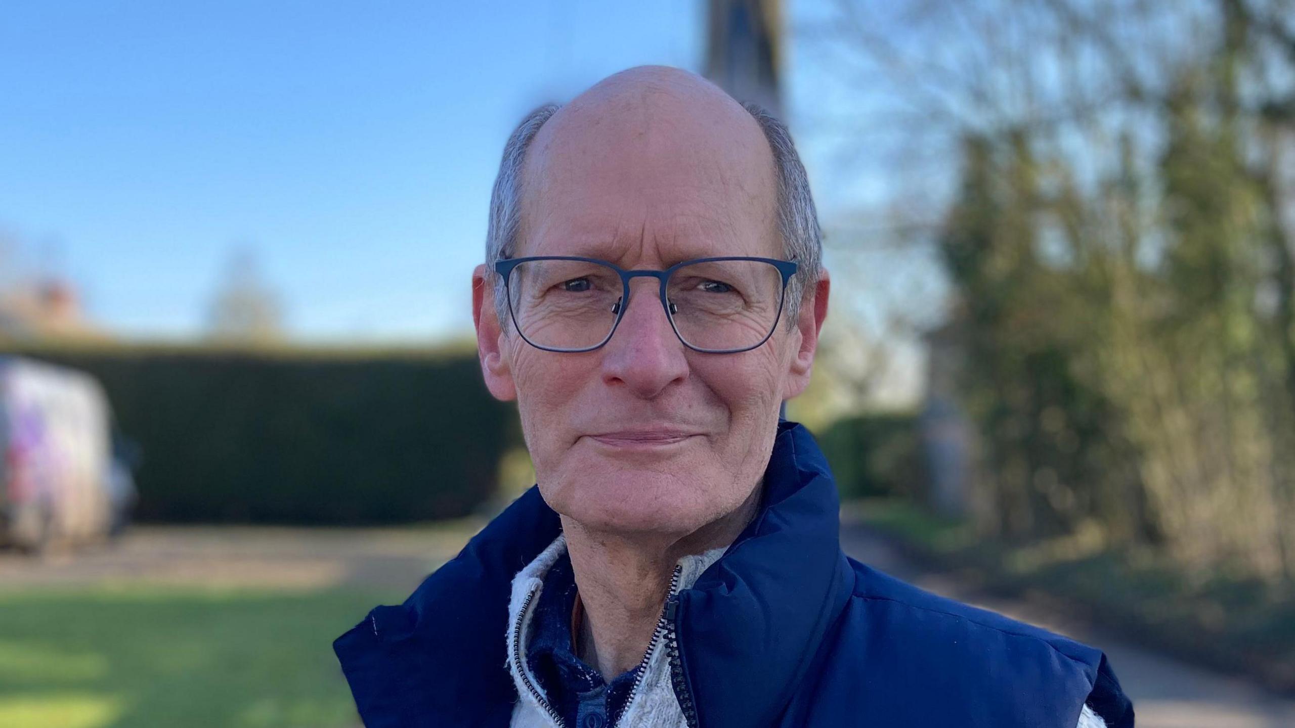 Churchwarden Richard Chatham smiles at the camera. He is largely bald with some grey hair around the side of his head. He wears a blue gilet with a white jumper underneath.