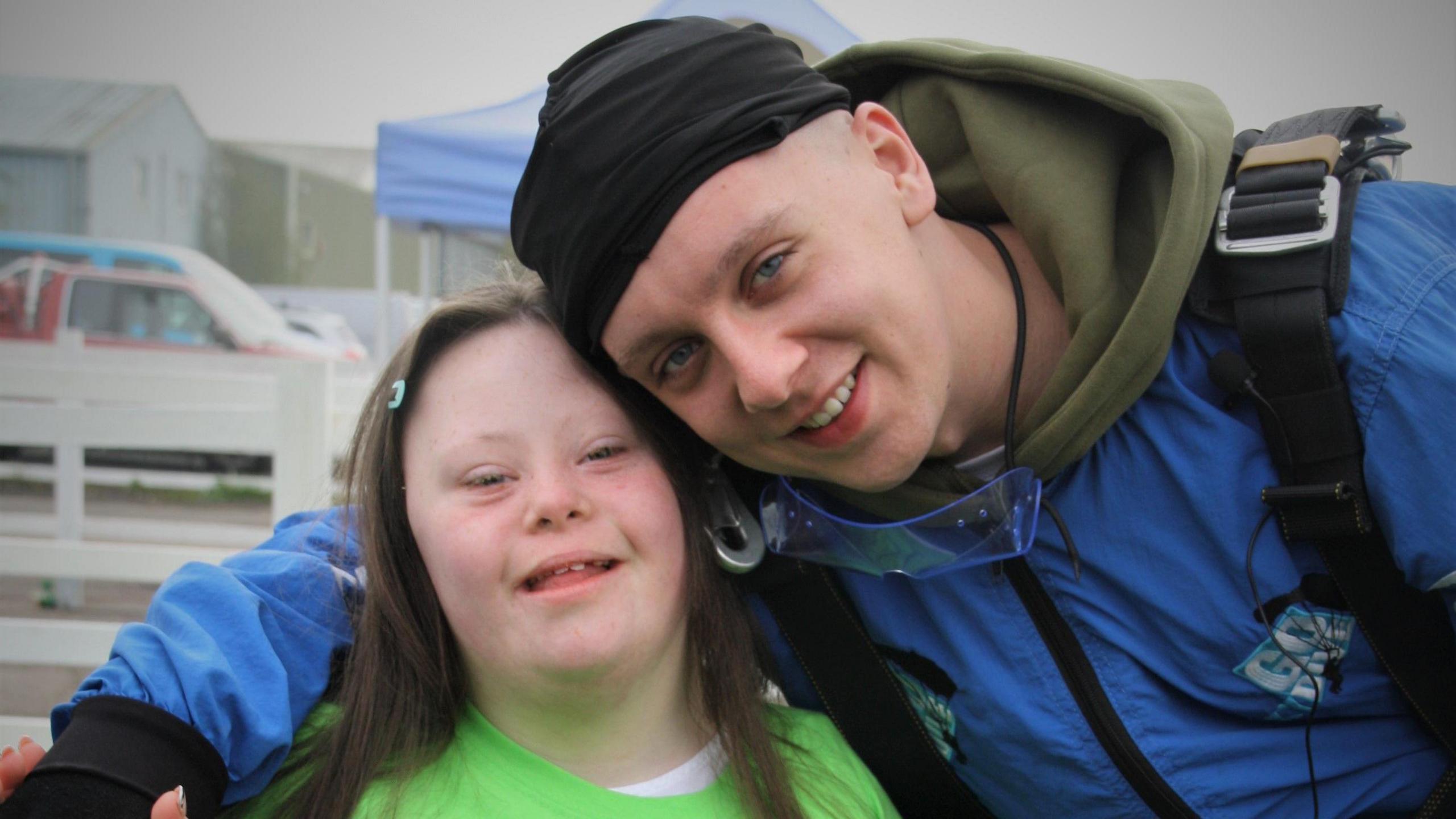 Gracie poses alongside Aitch, who has his arm around her. Gracie is wearing a bright green t-shirt. Aitch is wearing a blue skydiving jumpsuit. 
