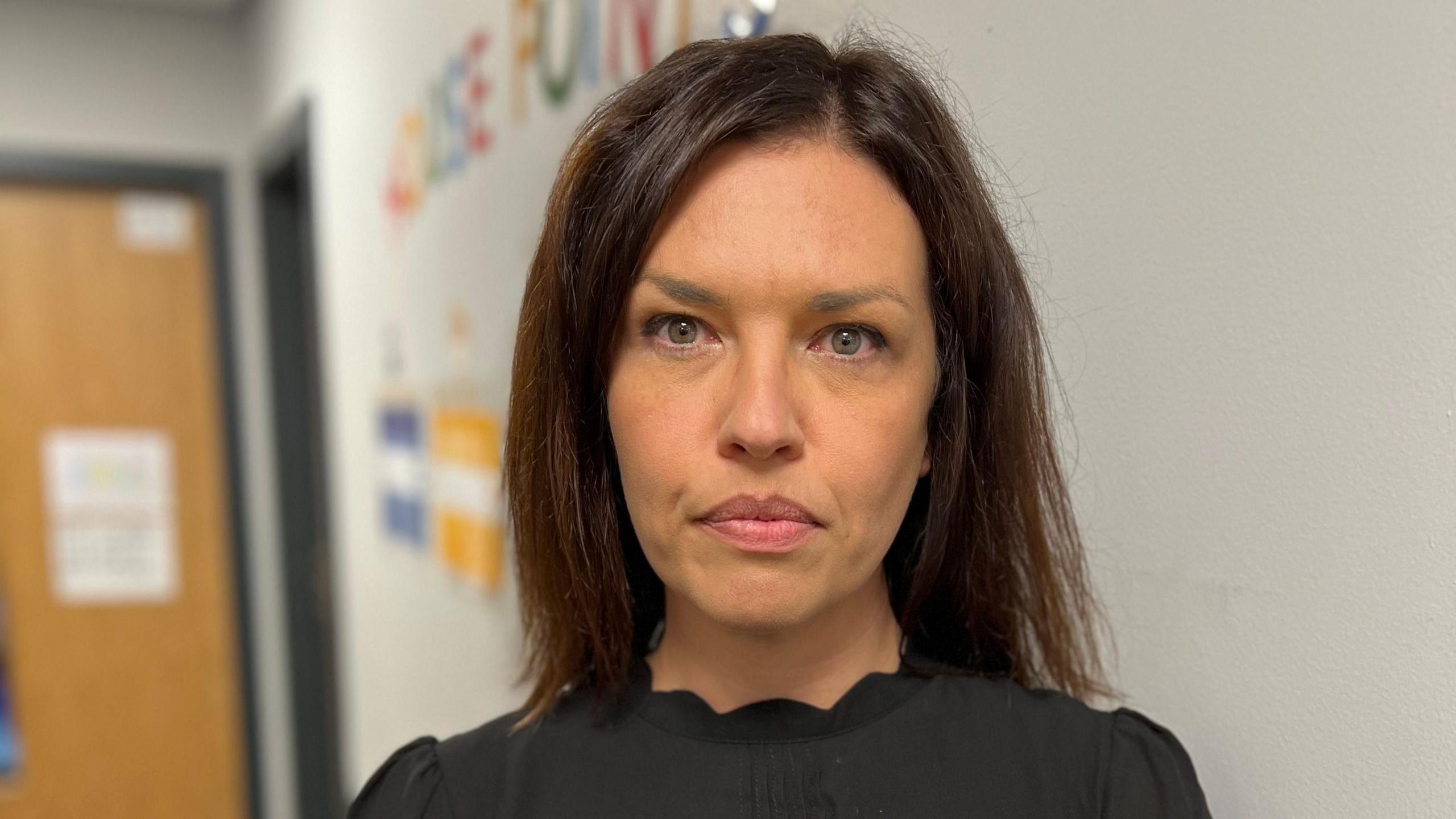 Michelle Catterson looks directly at the camera, she is wearing a black top and in the background a classroom wall can be seen with brightly coloured letters stuck on it