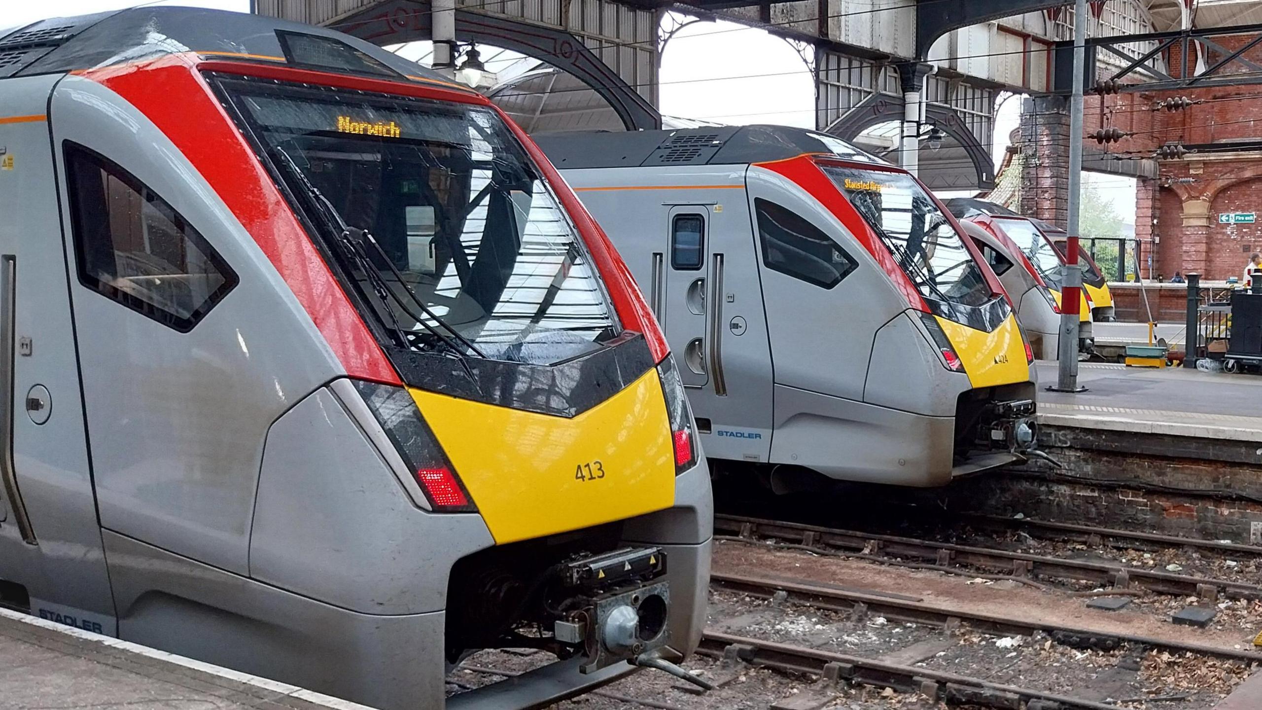 Four grey, yellow and red Greater Anglia trains sitting on parallel platforms inside a train station. The train nearest to the camera has 'Norwich' written in a display panel above its front windscreen. 